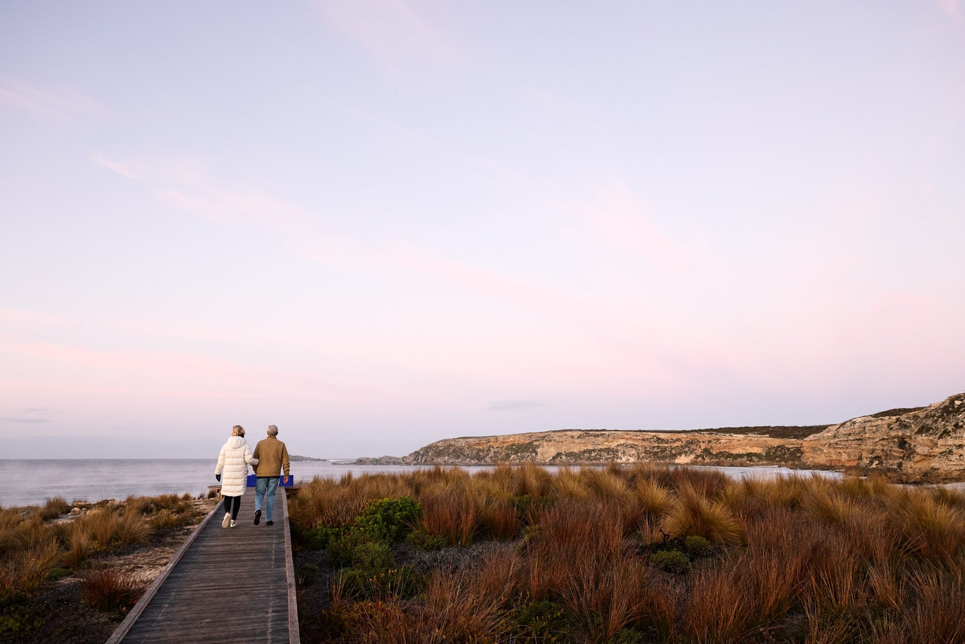 A couple walks on a boardwalk through a grassy landscape at sunset, with cliffs and a calm sea in the background.