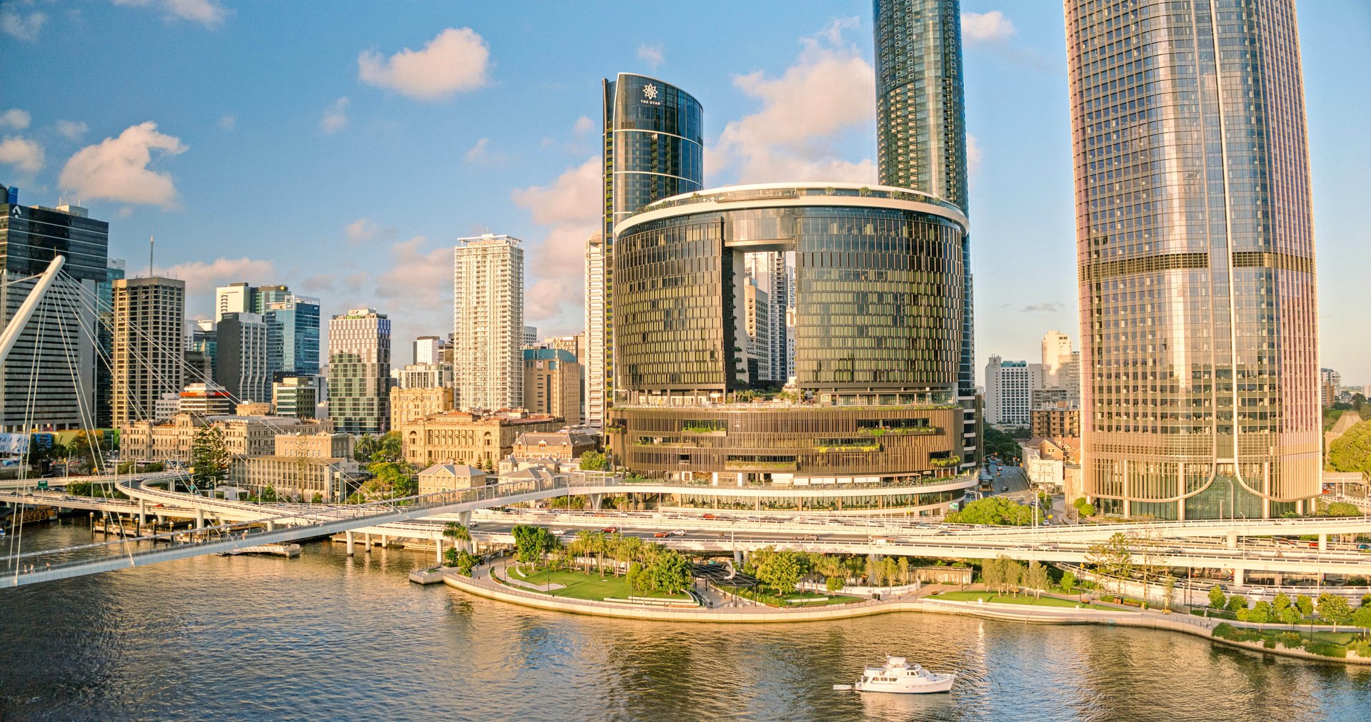 Aerial view of a modern cityscape with skyscrapers, a river, a bridge, and a boat on the water under a clear blue sky.
