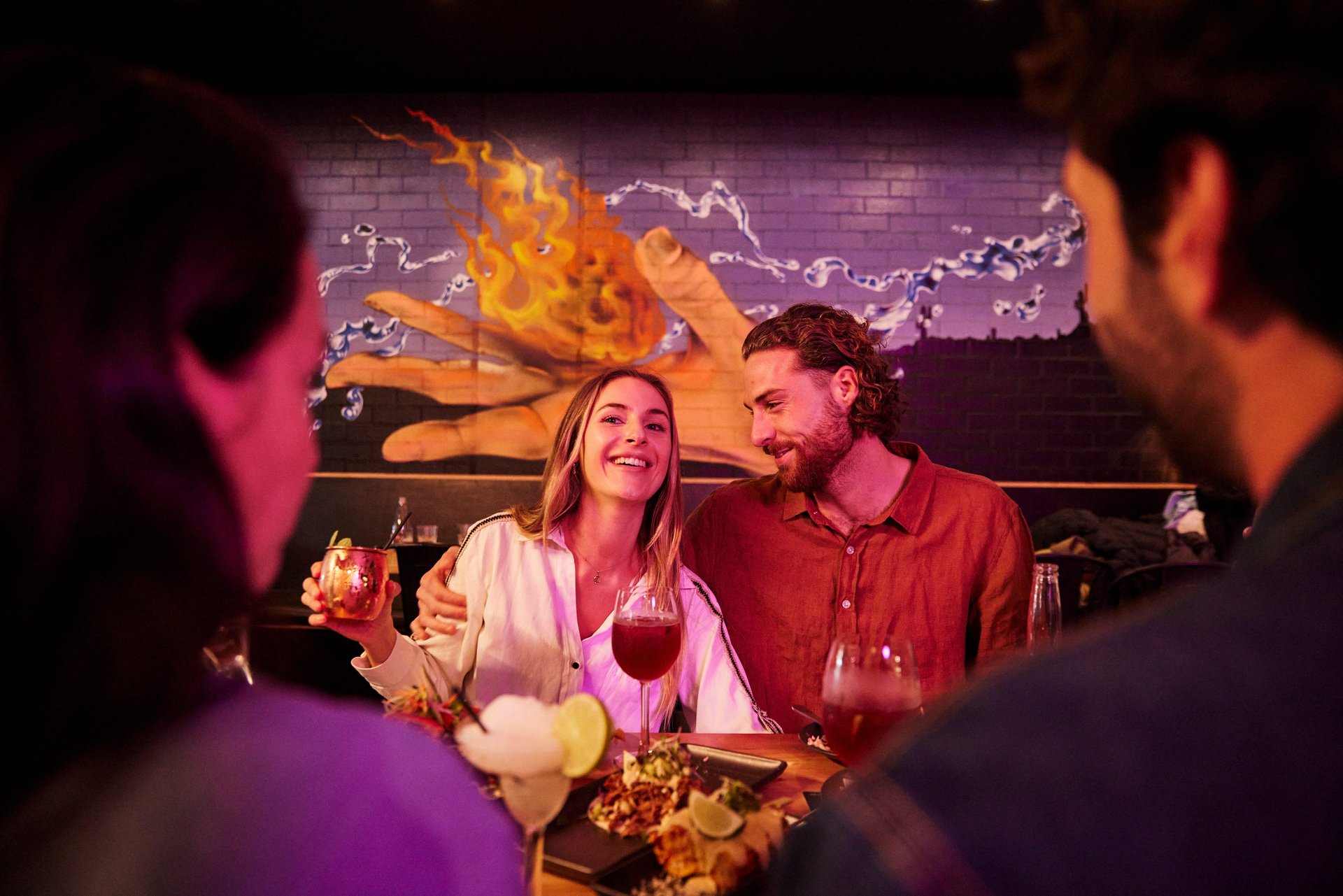Smiling couple with drinks at a lively bar; man has arm around woman, colorful mural of a flaming hand behind them.