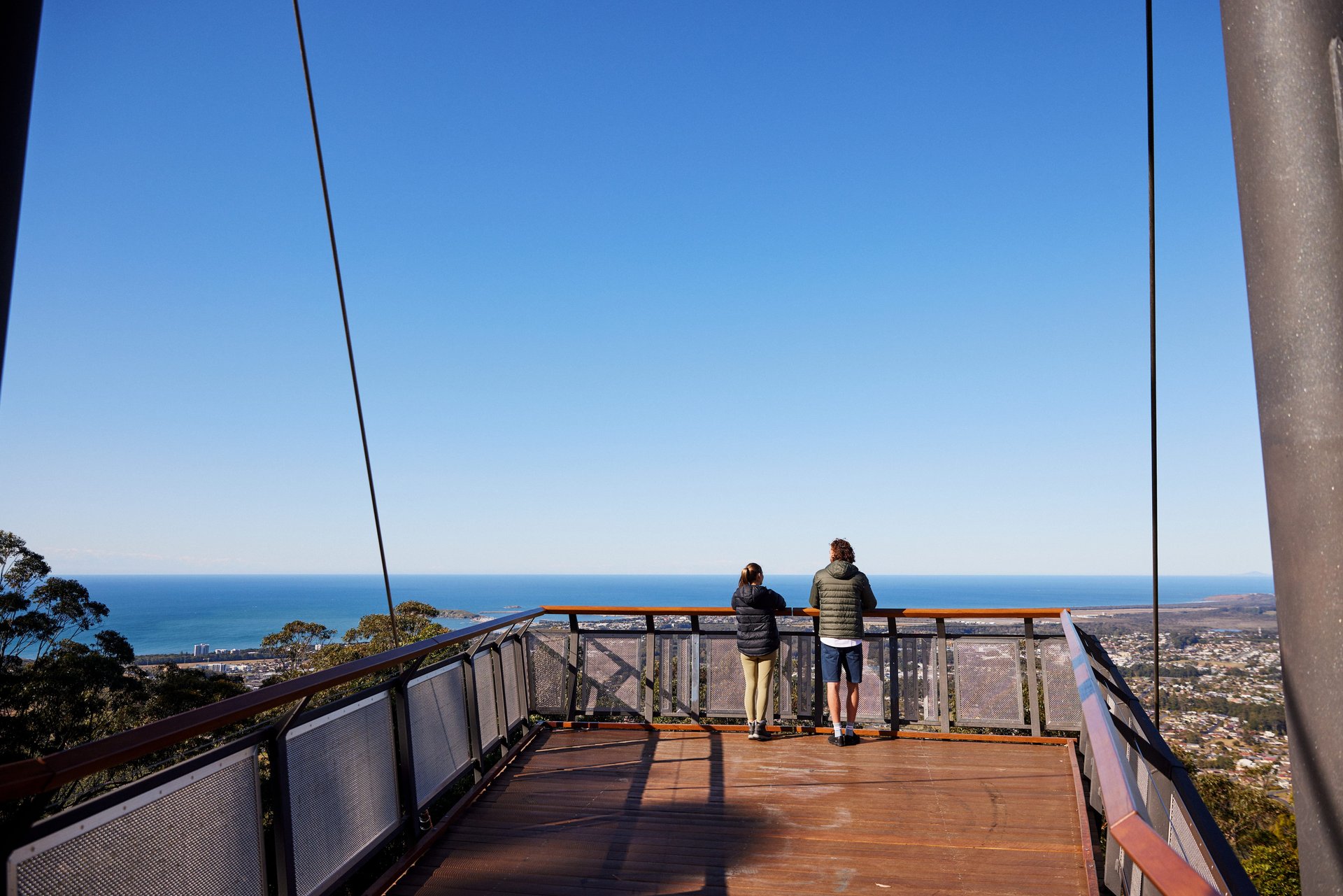 Two people in jackets stand at a wooden lookout deck, gazing over a coastal town and blue ocean under a clear sky.