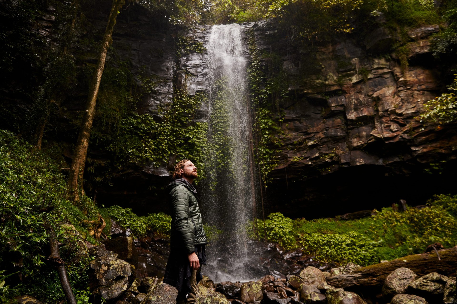 Person in green jacket standing on rocks, looking up at thin waterfall cascading down a mossy cliff in a forest.