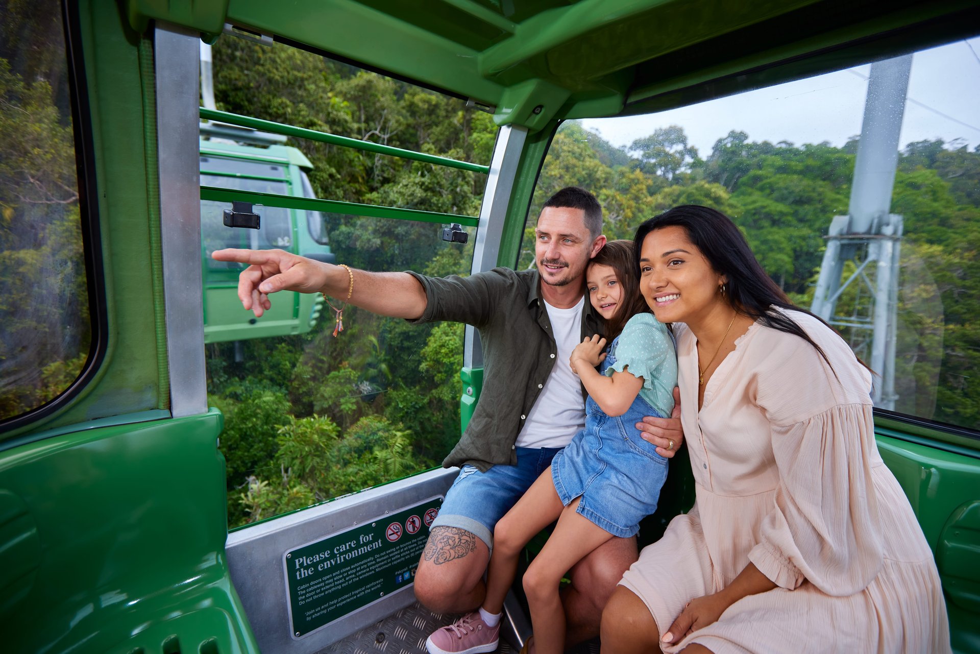 Family of three in green cable car above forest; father pointing out the window while mother and daughter smile.