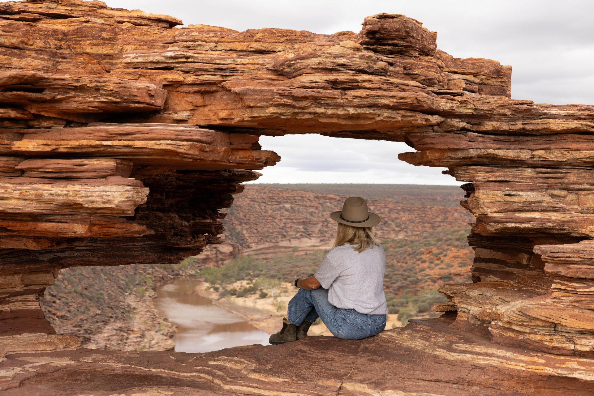 A person in a hat sits on a rocky ledge with a natural arch, overlooking a canyon and river under a cloudy sky.