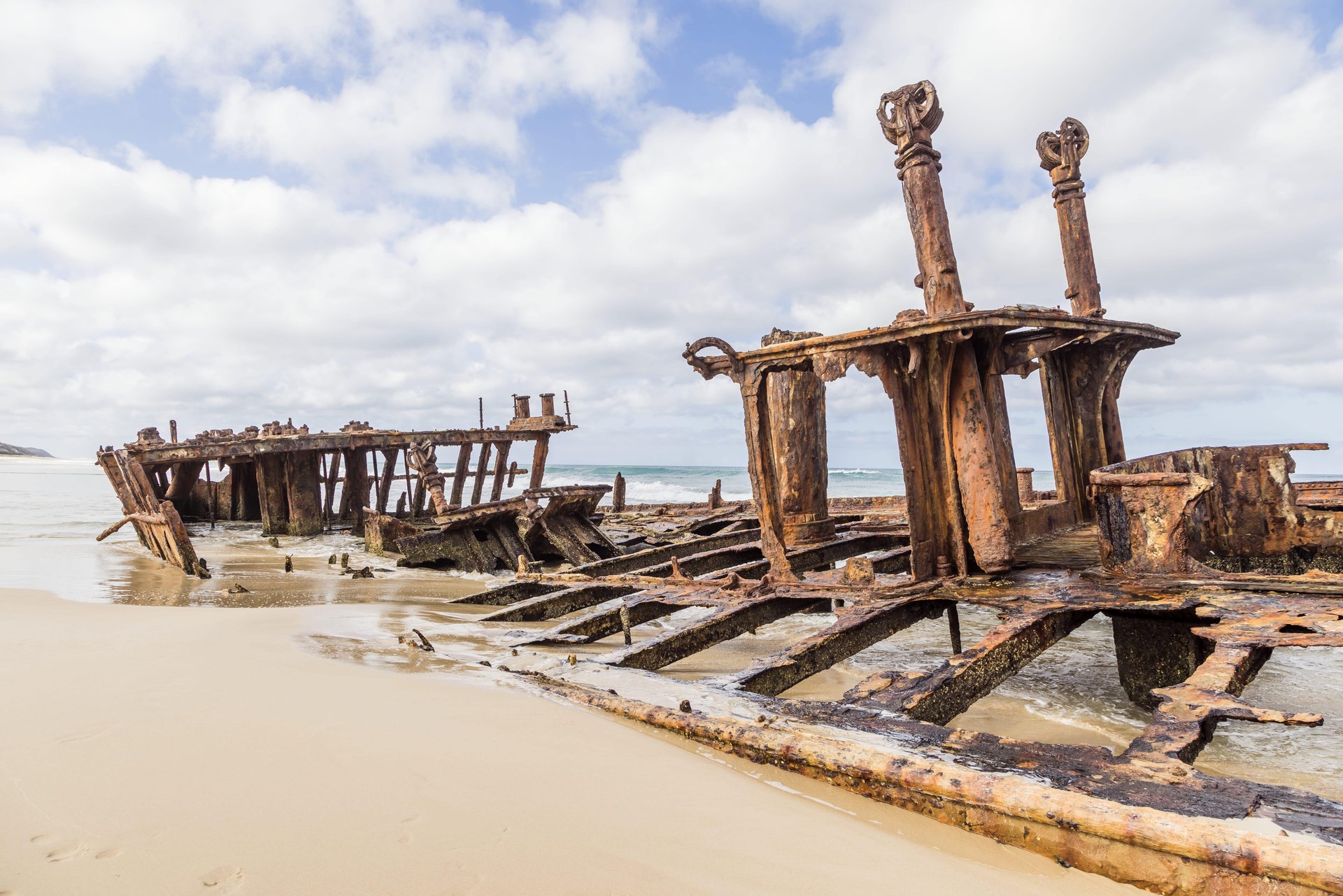 Rusty shipwreck remains partially buried in sand on a beach, with a cloudy sky and ocean waves in the background.