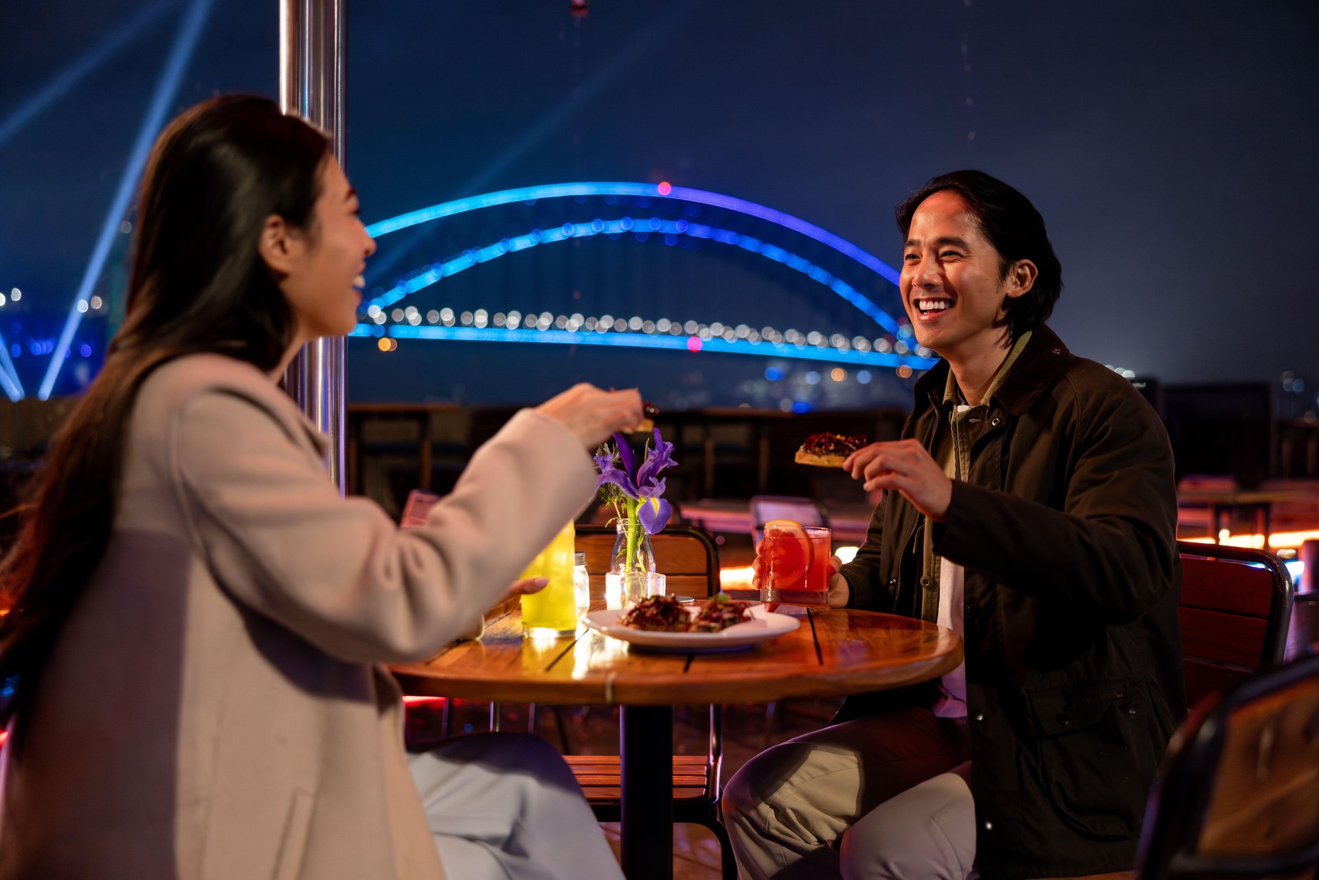 Two people enjoying a meal at an outdoor restaurant at night, with a lit-up bridge in the background.