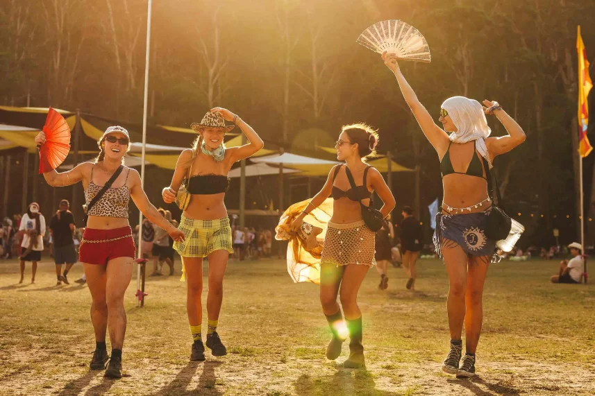 Four people in festival attire walk joyfully on a sunny day, holding fans. Tents and trees are visible in the background.