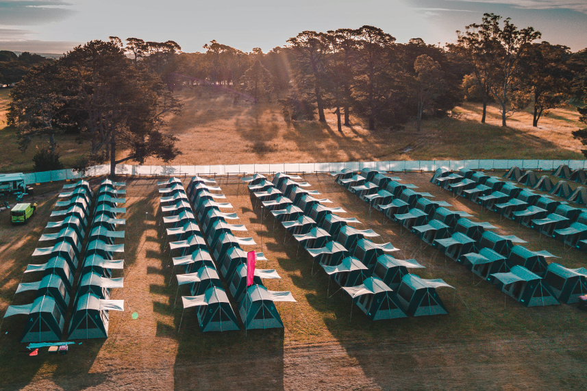 Aerial view of a campsite with rows of green tents on grassy terrain, surrounded by trees under a clear sky.