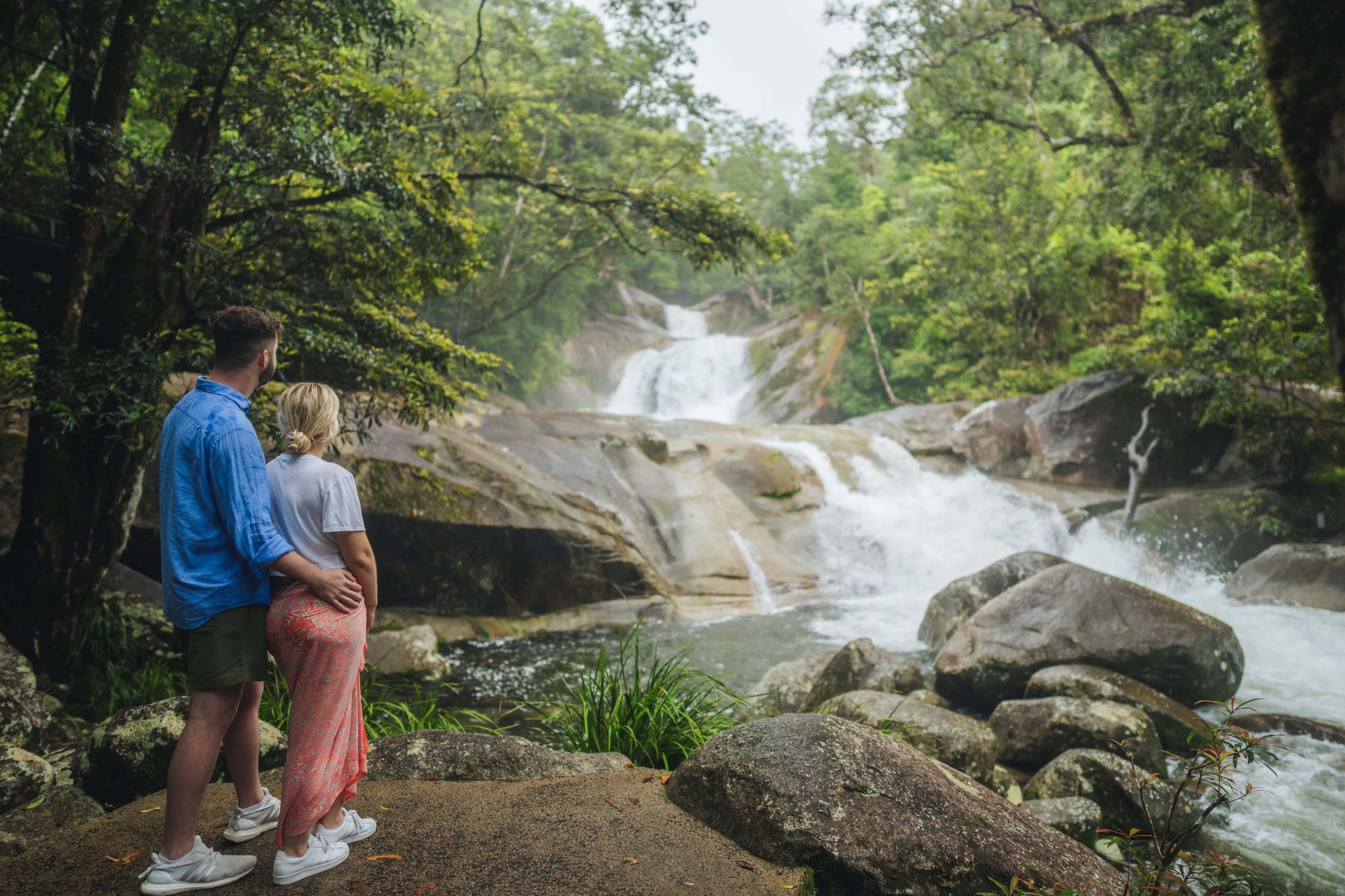 Couple standing on rocks with arms around each other, gazing at a cascading multi-tier waterfall in a lush green forest.