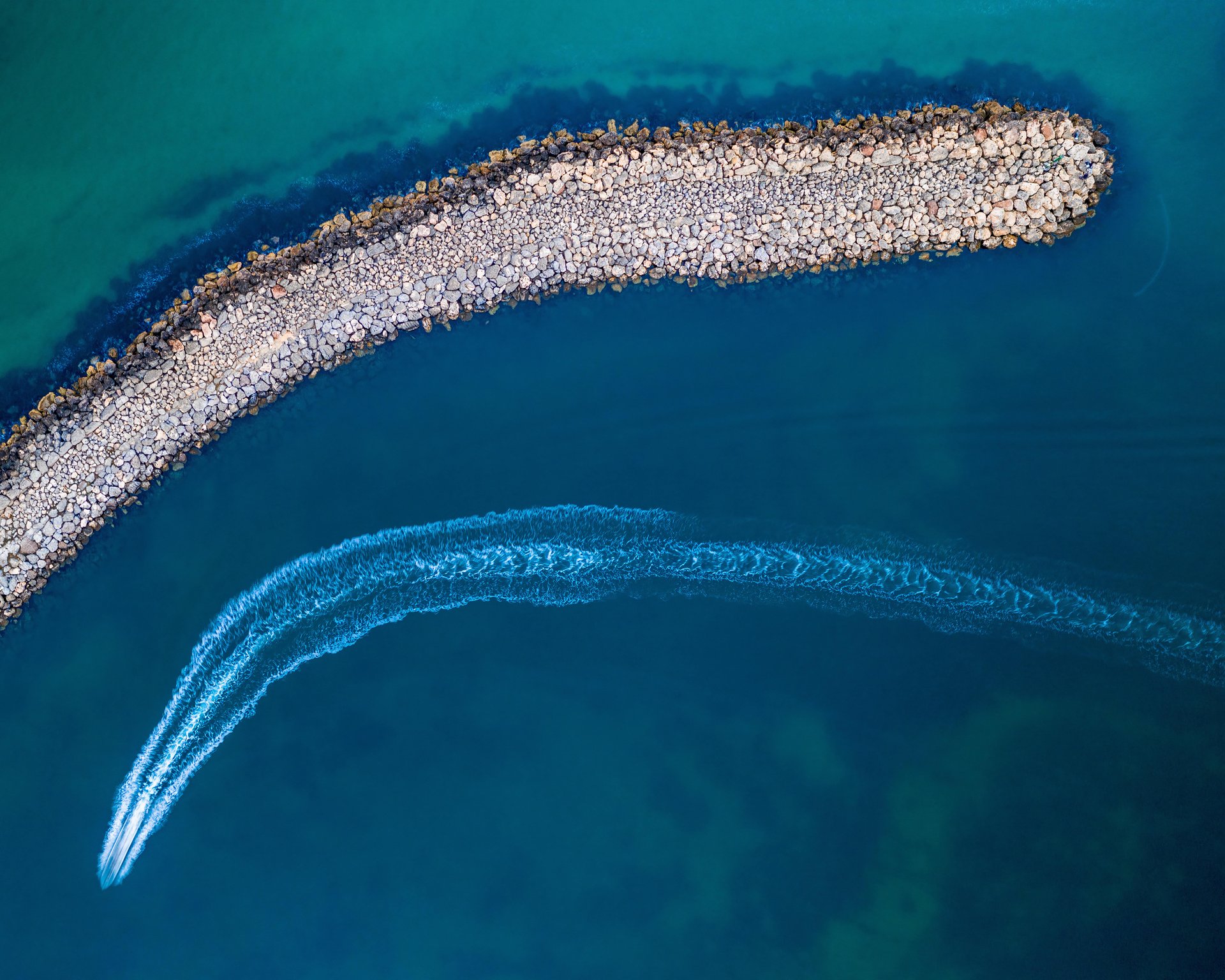 Aerial view of a speedboat creating a curved wake near a long, rocky breakwater in clear blue-green water.