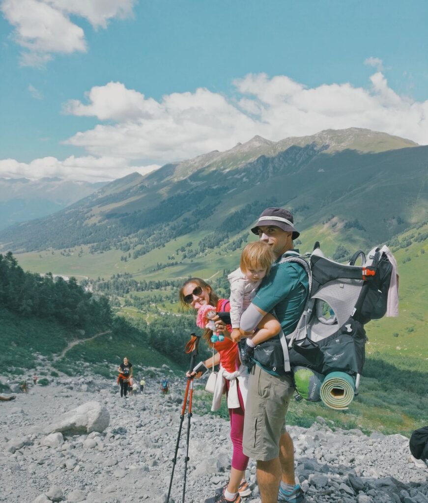 Eine Familie wandert auf einem felsigen Pfad in den Bergen, mit einem Kind in einer Trage und Rucksäcken, umgeben von grünen Hügeln und blauem Himmel.