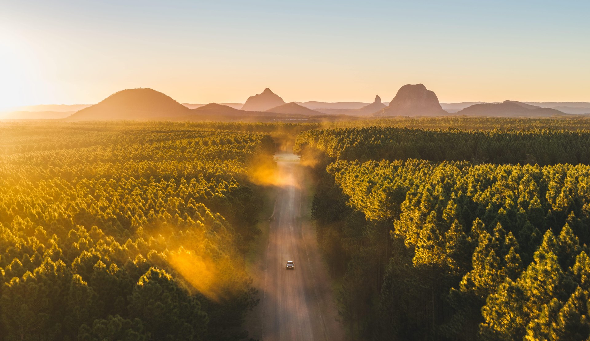 Aerial view of a car driving on a dirt road through a forest at sunset, with distant mountains silhouetted against the sky.