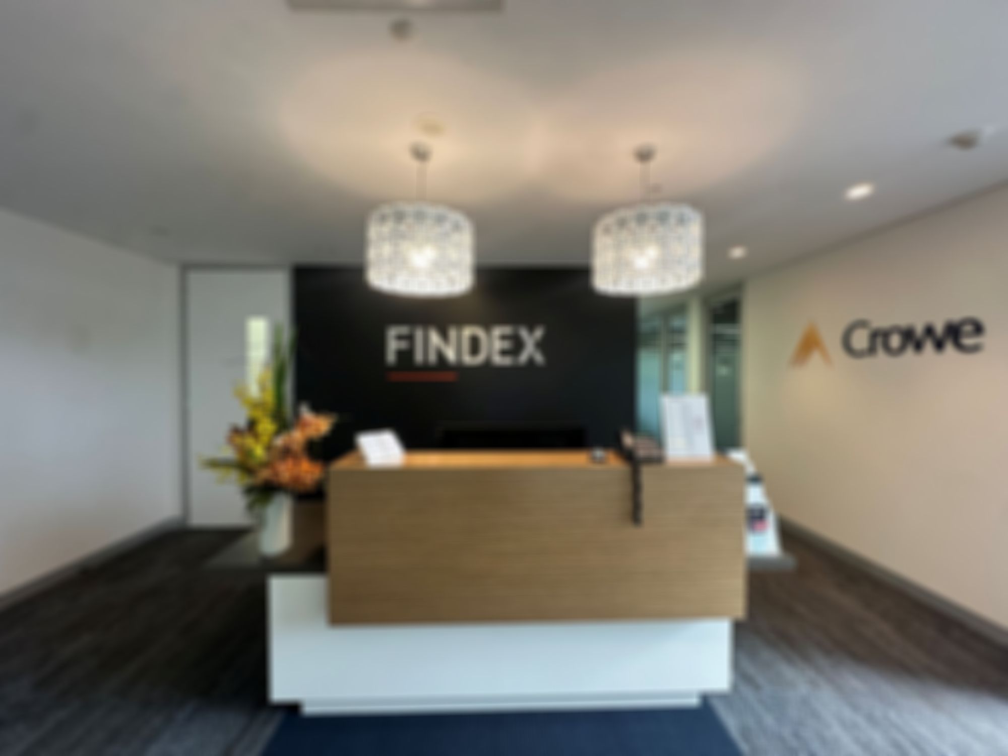 Findex Norwest reception area with a wooden desk, floral arrangement, decorative hanging lights, and signs reading "FINDEX" and "Crowe" on the wall.