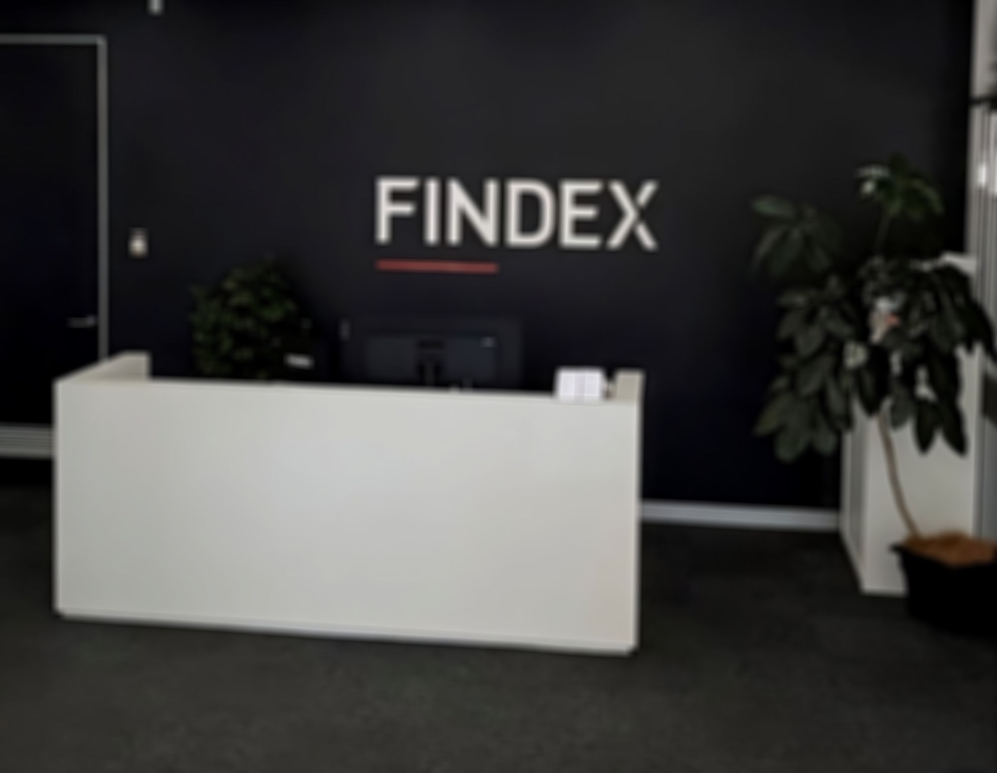 Findex Ballina office reception area with a white desk, computer, potted plants, and "FINDEX" logo on a dark wall.