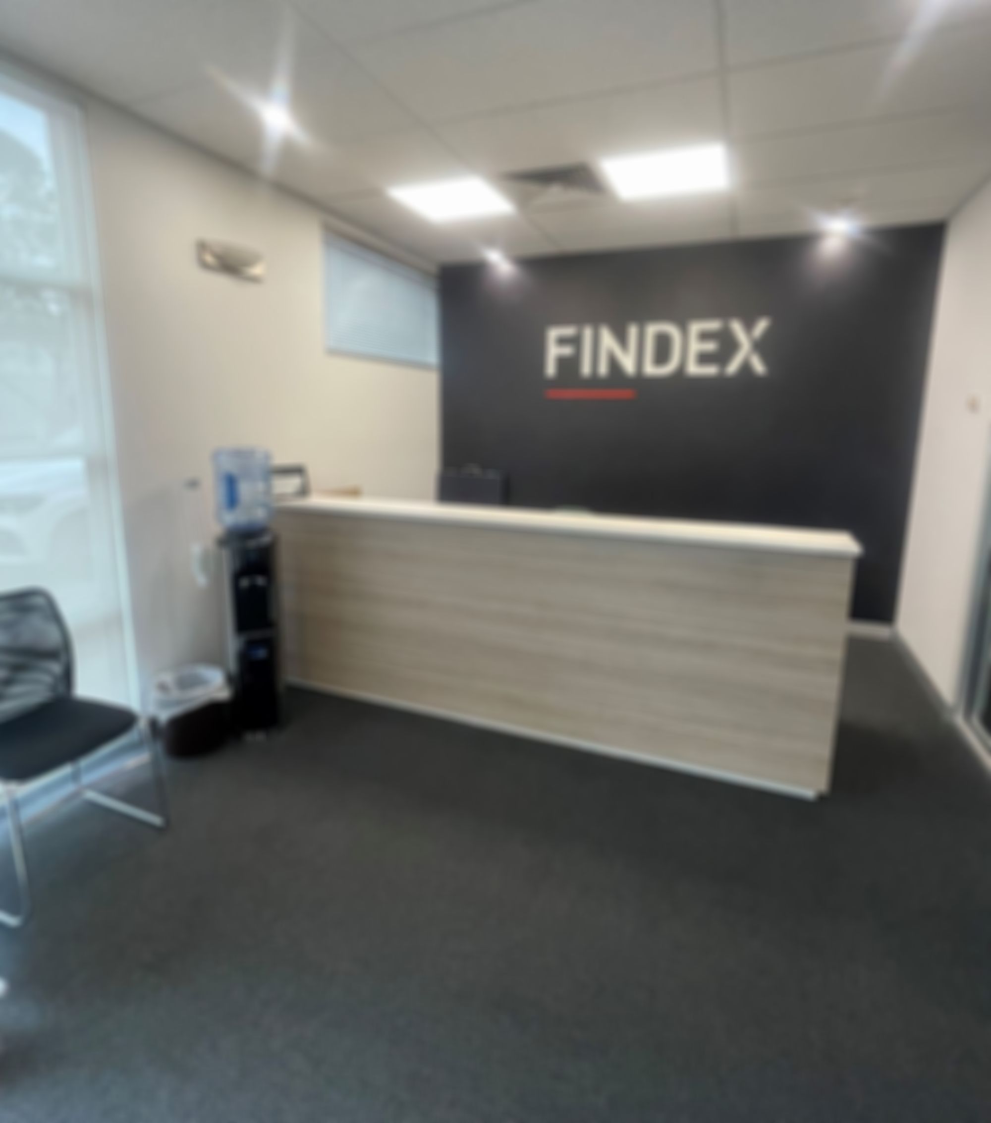 The Findex Traralgon office reception area with a wood-accented desk, "FINDEX" sign, water cooler, black chairs, and soft lighting.