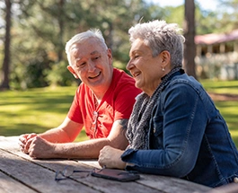 Older couple smiling and talking while sitting at a picnic table in a sunny park, surrounded by trees.