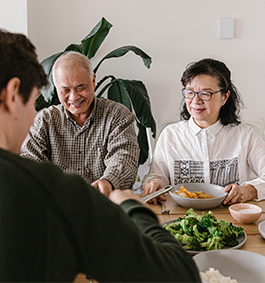 Three people sit around a table sharing a meal, with bowls of vegetables and rice. Two are smiling, and a plant is visible in the background.