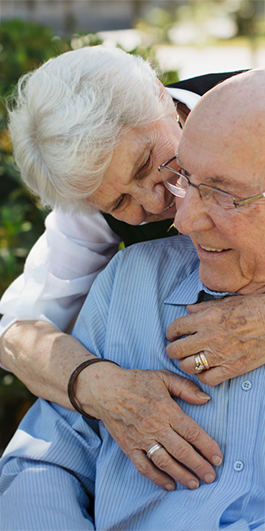 Elderly woman affectionately hugs an elderly man from behind, both smiling, outdoors with greenery in the background.