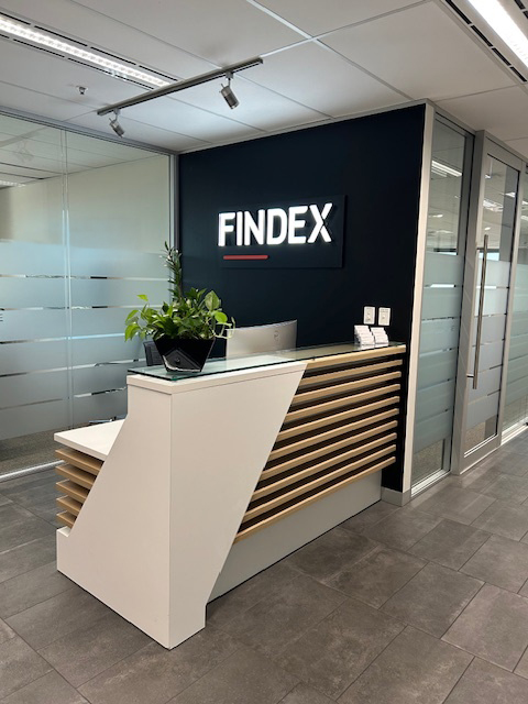 Findex Adelaide reception desk. Desk has computer monitor, green plant on top of desk and contact cards. Large Findex sign is next to the desk.