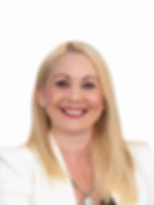 Smiling woman with long blonde hair and a necklace, wearing a white blazer against a plain background.