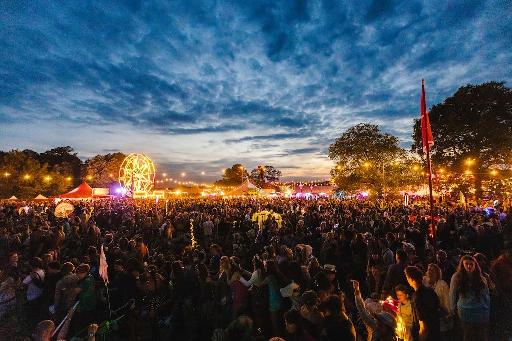 Large crowd gathered at Shambala Festival during dusk, with an illuminated Ferris wheel visible in the distance, colorful stage lights, red tents, and trees silhouetted against a dramatic blue and orange twilight sky