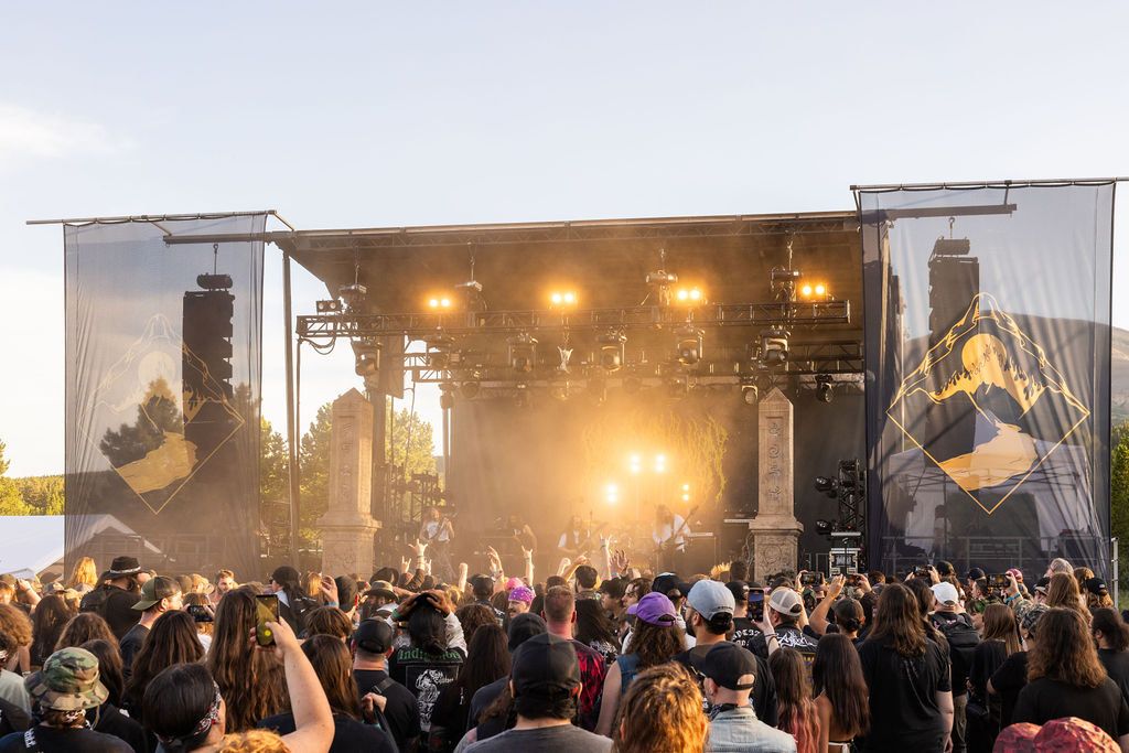 Outdoor music festival stage at golden hour with bright stage lights illuminating performers, surrounded by a large crowd of attendees. Professional lighting rig and speaker stacks are visible on the stage structure, with branded banners featuring mountain diamond logos hanging on either side. Trees and sky visible in the background creating a sunset atmosphere.
