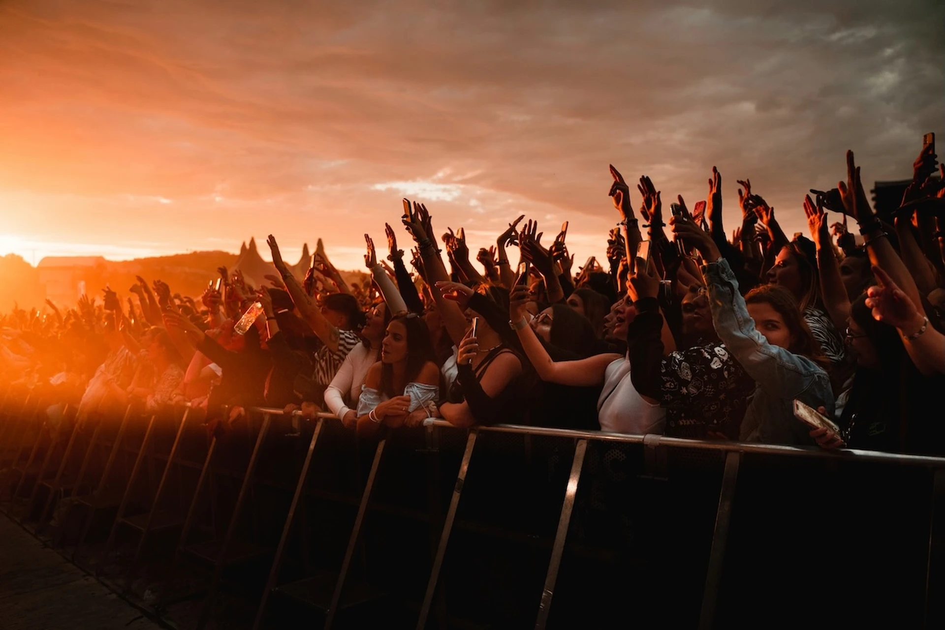 Concert crowd with raised hands at an outdoor event during sunset, creating a silhouette effect under an orange sky.