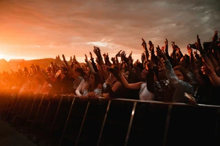 Concert crowd with raised hands at an outdoor event during sunset, creating a silhouette effect under an orange sky.