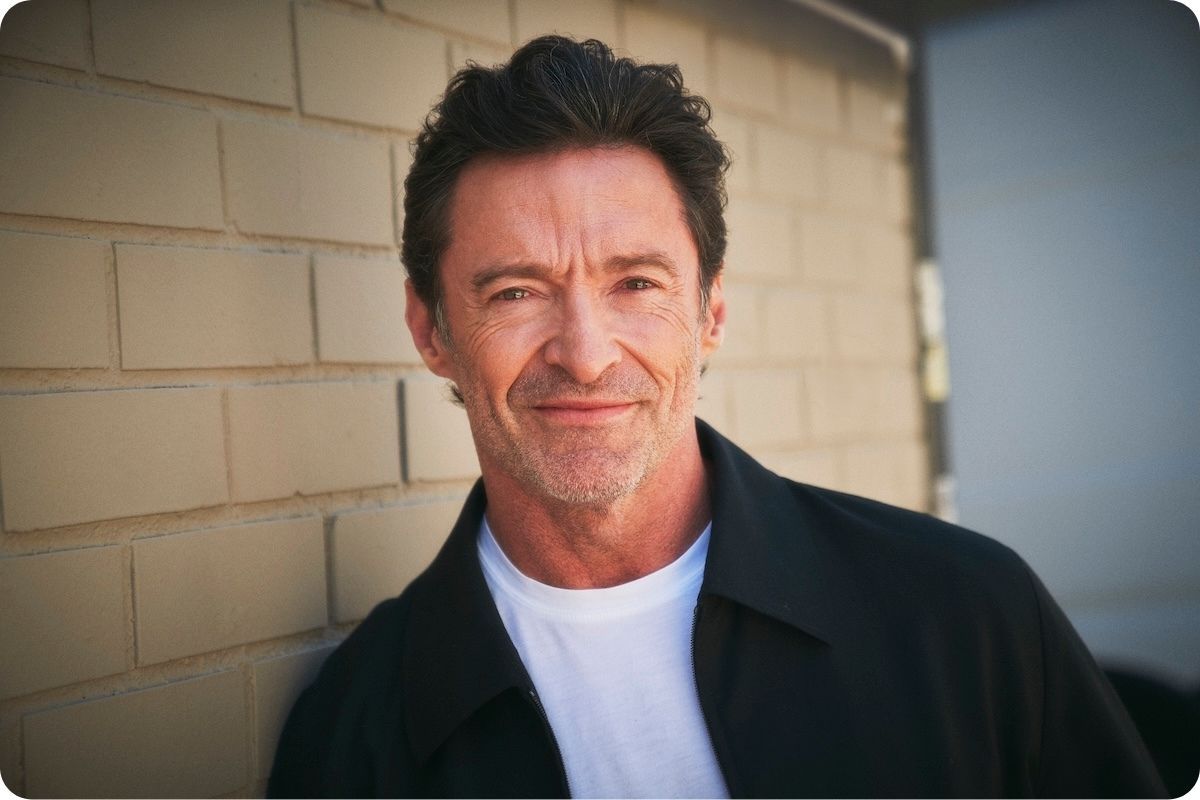 Hugh Jackman smiling to camera with a white t-shirt and black jacket.