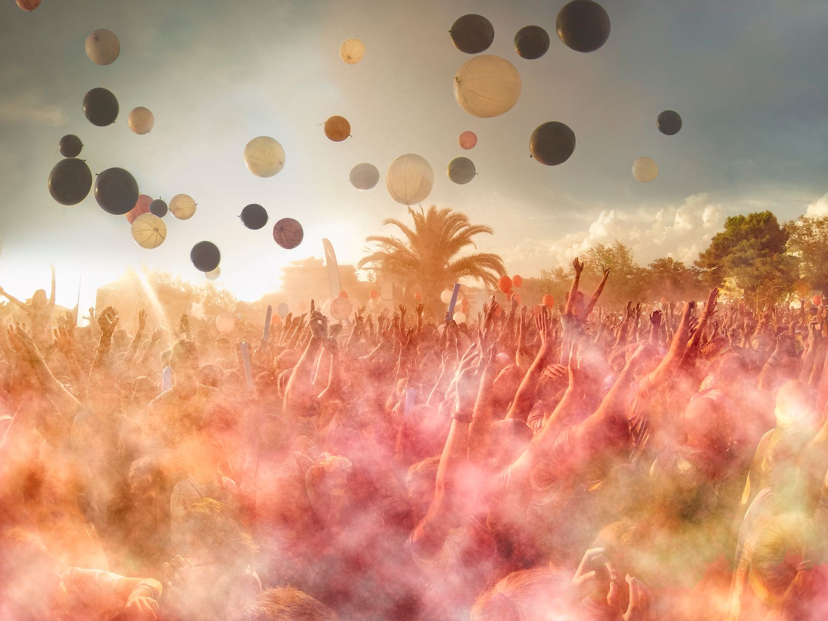 A very colourful photo of a vibrant festival, with hundreds of patrons with their hands in the air. They have thrown colourful dust into the air, so that there is a misty overlay of yellow, orange and pink. The sun is setting in the background, and there a colourful balloons also pictured in the sky.