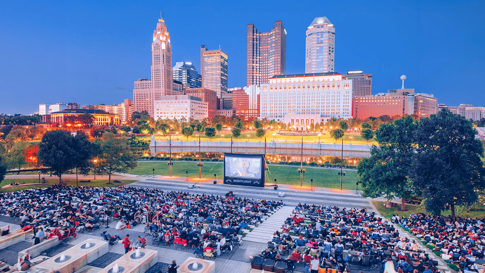 An aerial view of a large outdoor cinema event at dusk, with hundreds of people seated in front of a NightLight screen against a glowing city skyline.