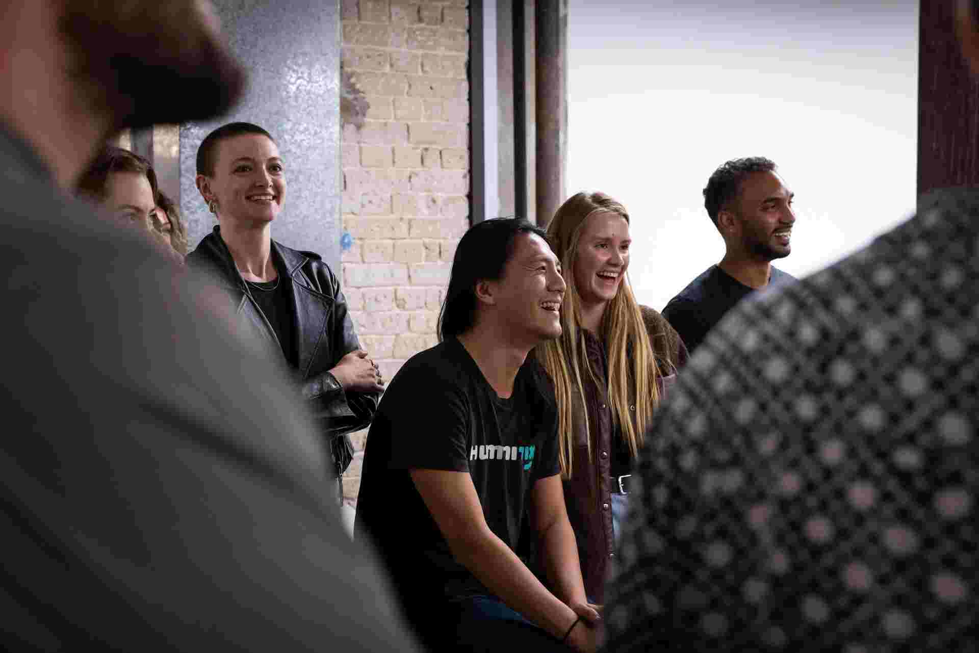 A bokeh style photo of multiple Humanitix staff smiling and laughing in the Humanitix office, listening to an off-picture speaker present.