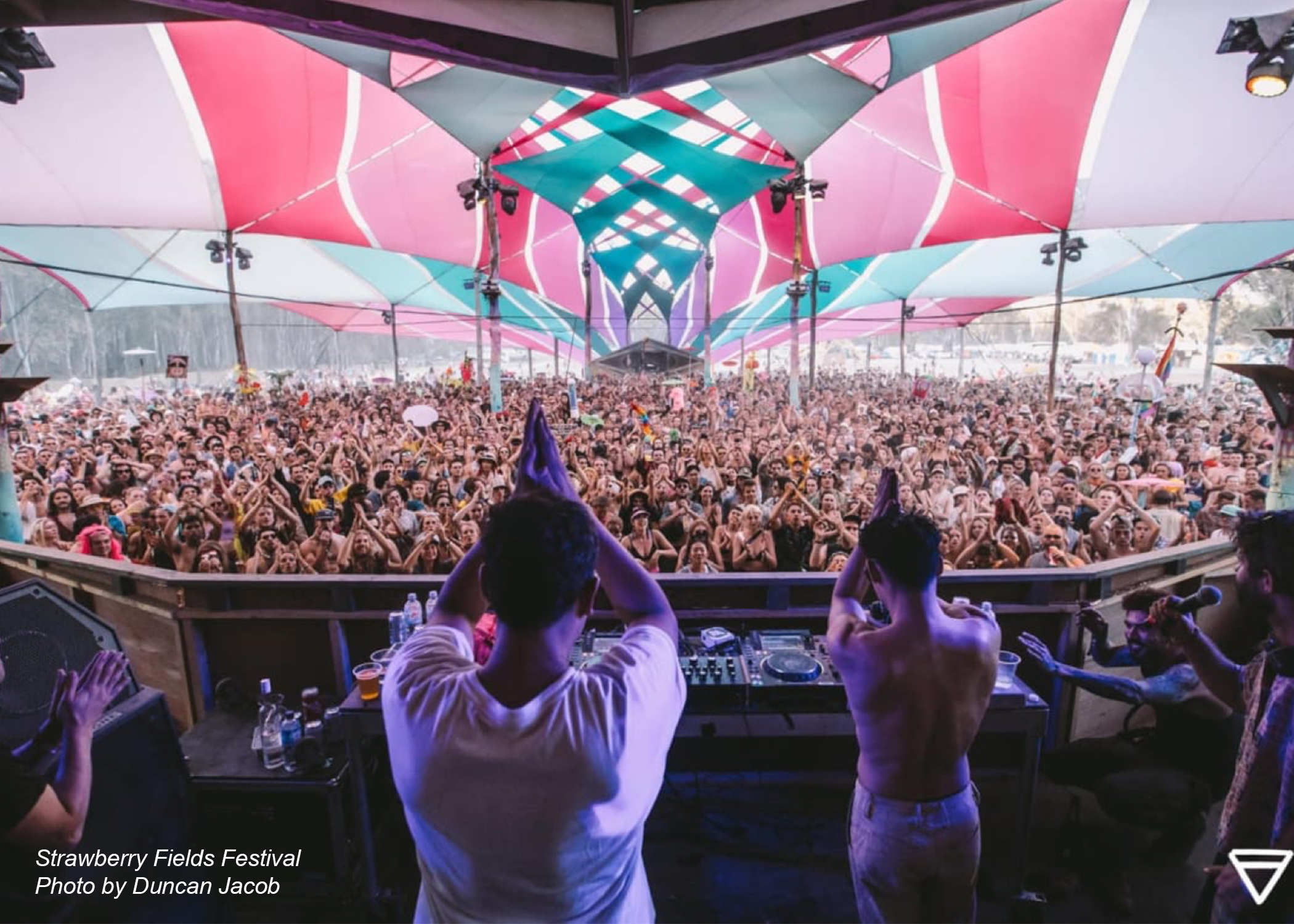 A photo taken from behind two DJs playing to a large crowd of attendees at the Strawberry Fields Music Festival. They have their hands up in the air in a 'prayer' shape, thanking the crowd. There are colourful shade cloths above the crowd, adding colour and vibrancy to the event atmosphere.