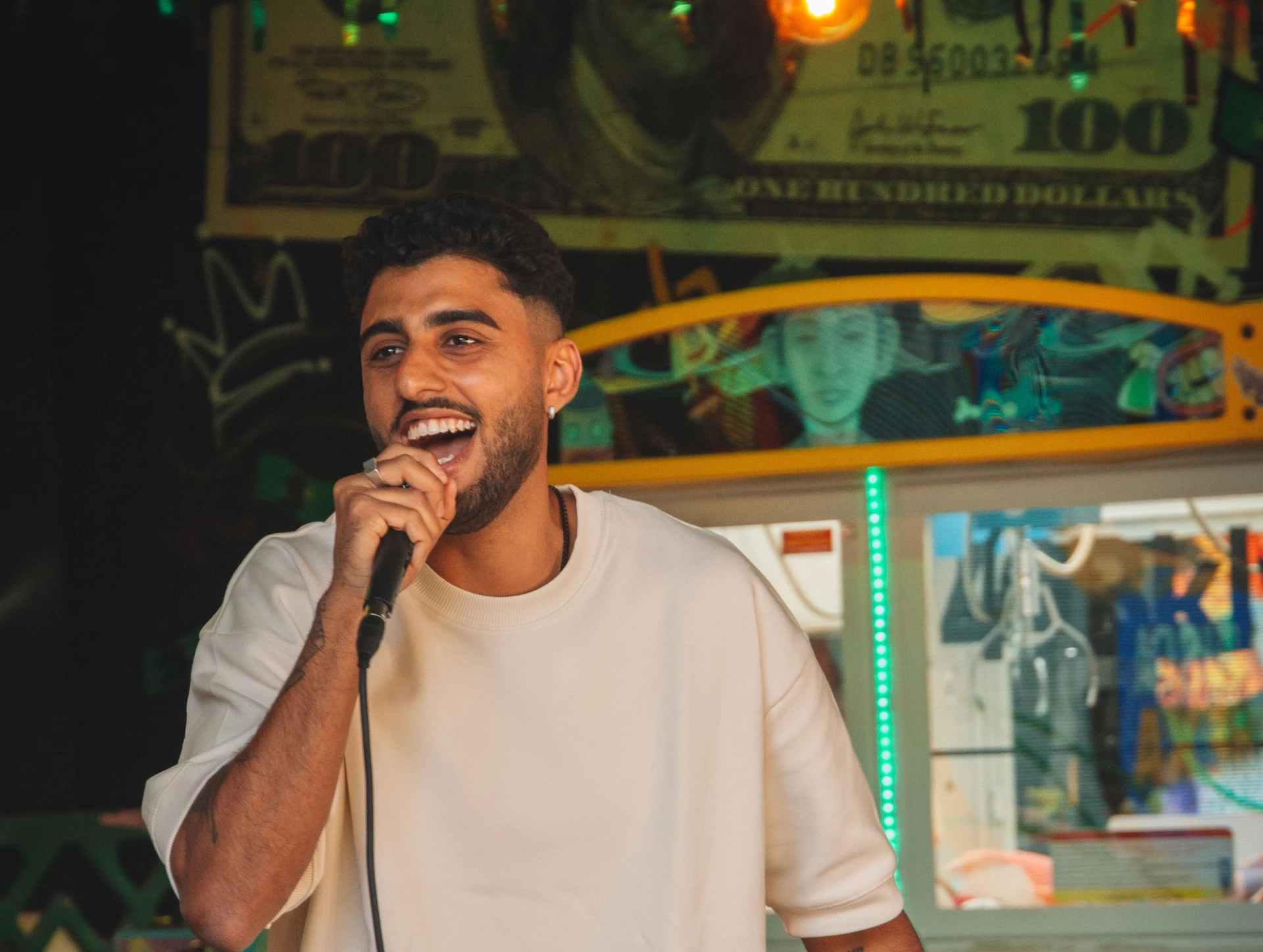 A smiling man in an oversized cream t-shirt holds a microphone at a lively indoor venue decorated with gold tinsel, neon green lights, and a large novelty banknote on the wall behind him.