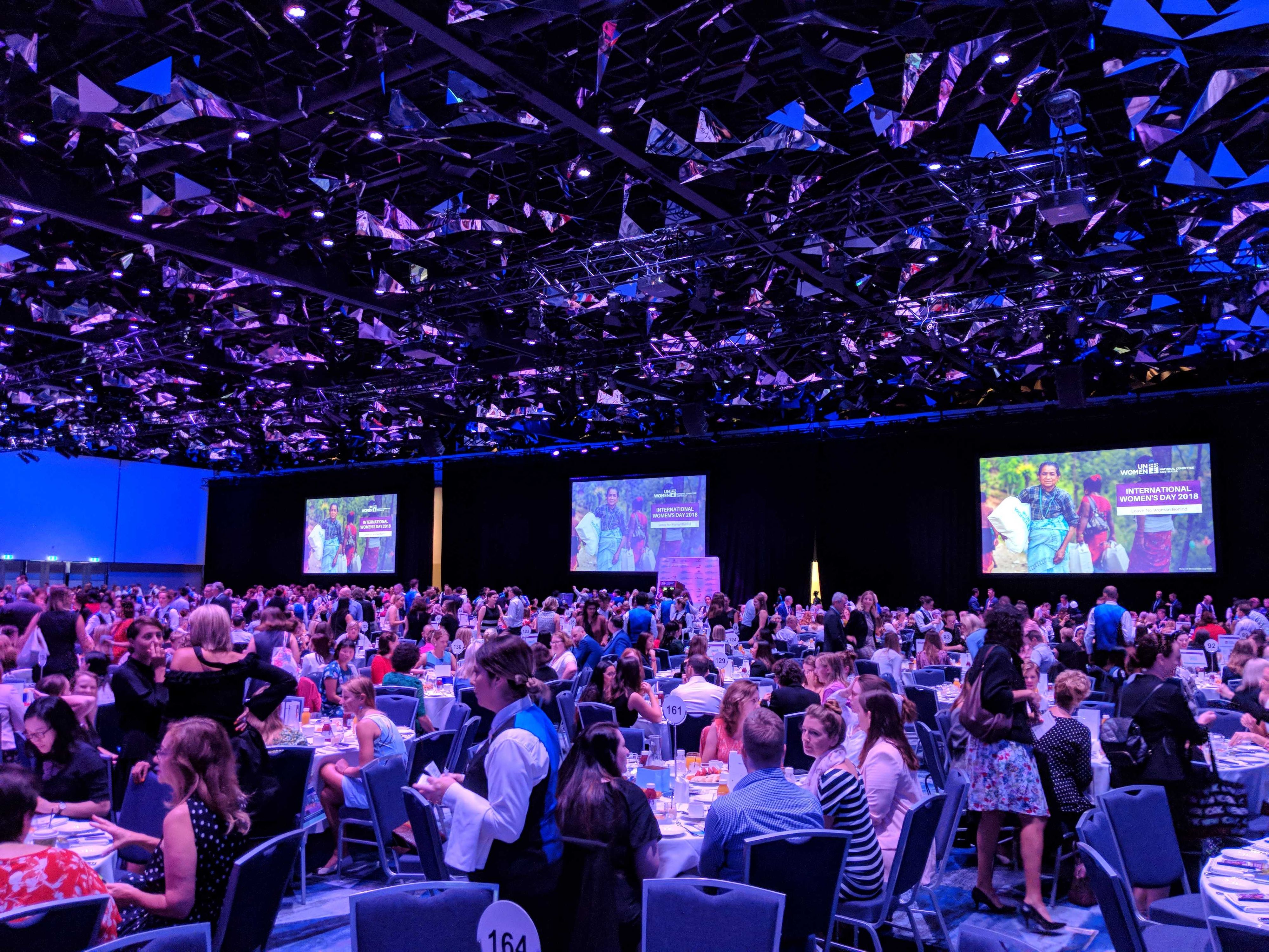 A picture of the large room, filled with tables in a gala style event. There is purple overhead lighting giving a professional look to the event, with patrons seated at their tables chatting. Screens of projected presentation slides are on the back wall of the room.