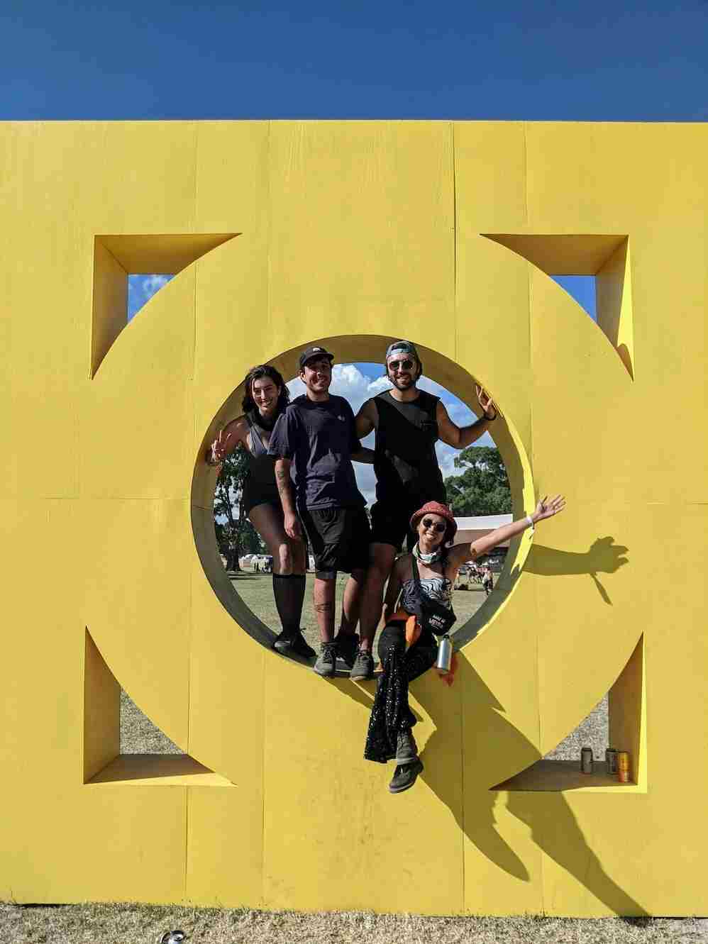 A picture of 4 Humanitix staff standing and crawling within a large, yellow artistic structure that features at a music festival. The structure has a hole in the centre where the staff have climbed into, and contains gaps in each corner of the rectangular structure that peers through to the open field it is placed in.