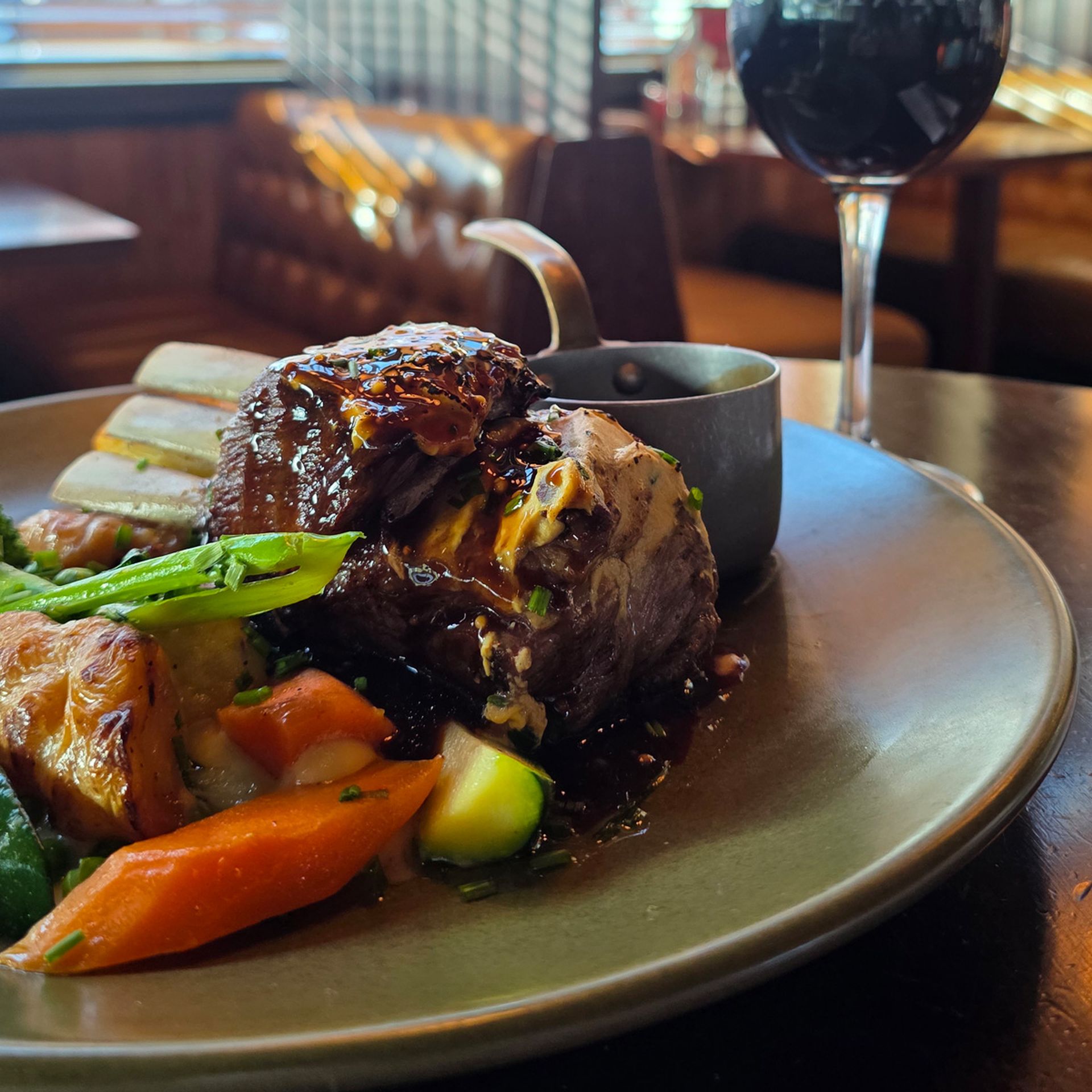 A gourmet dish with lamb, vegetables, and sauce on a plate, accompanied by a glass of red wine in a cozy restaurant setting.
