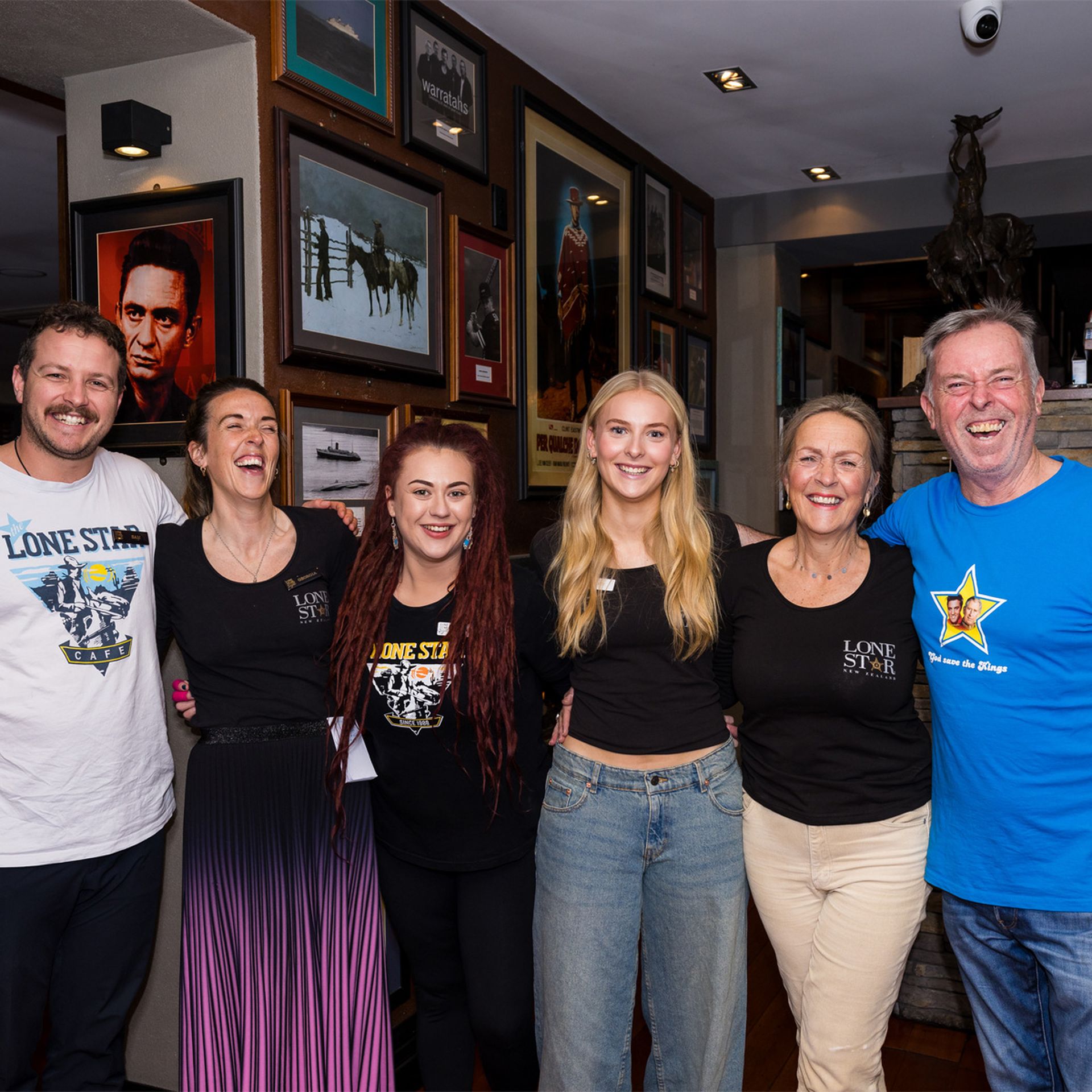 A group of six people smiling, standing close together in a room with framed pictures on the walls. They appear to be enjoying a casual gathering.