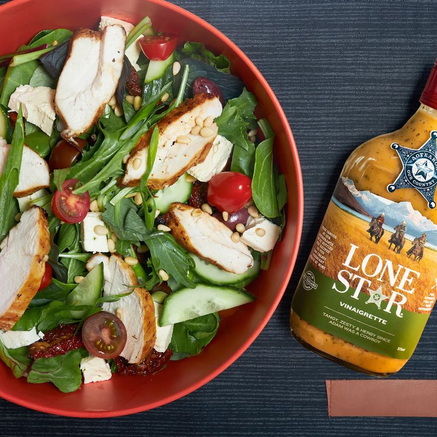 A salad with grilled chicken, cherry tomatoes, cucumbers, and greens in a red bowl next to a bottle of Lone Star vinaigrette dressing.