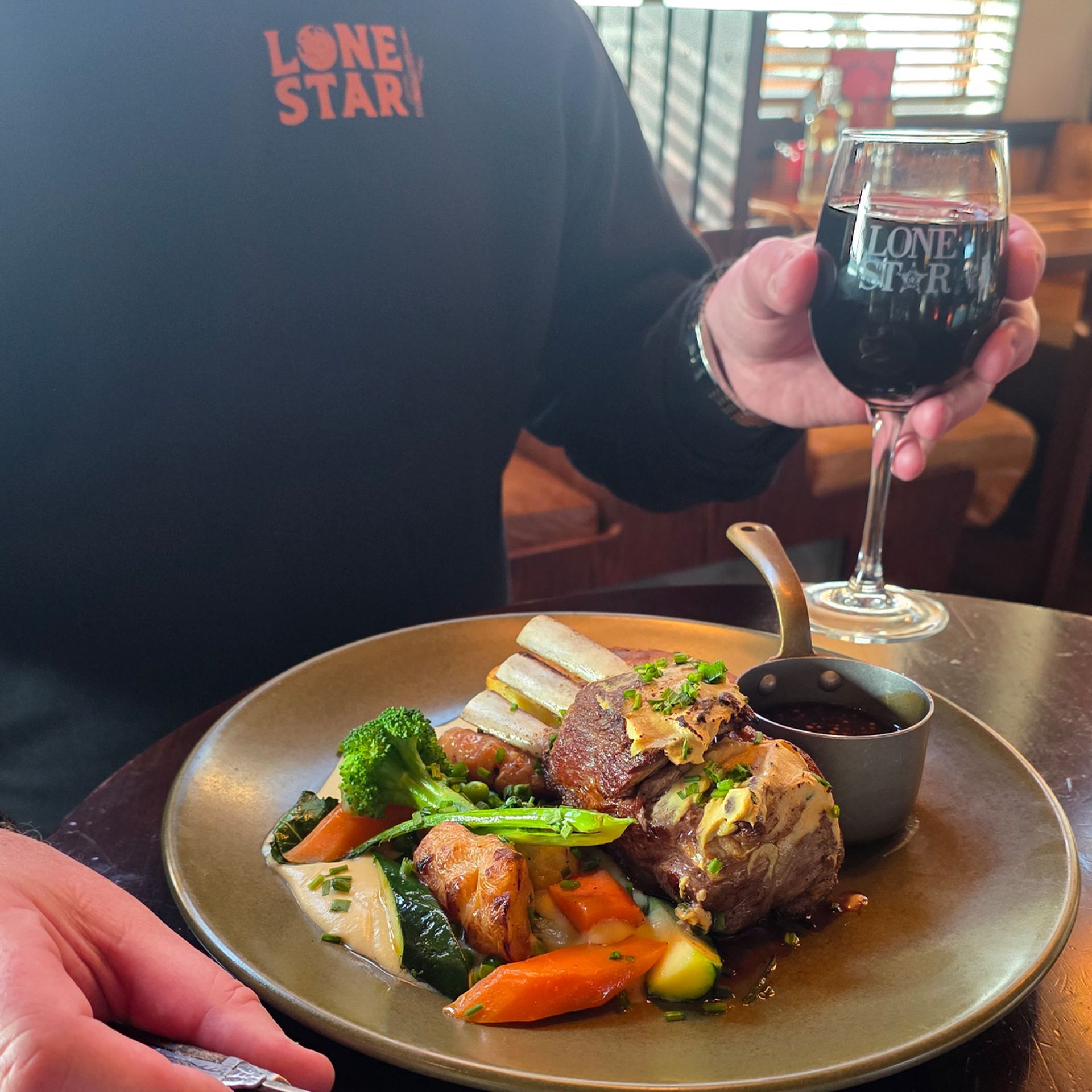 Person holding a glass of red wine, with a High Noon Lamb Shoulder Rack, and sauce on a table at a restaurant.