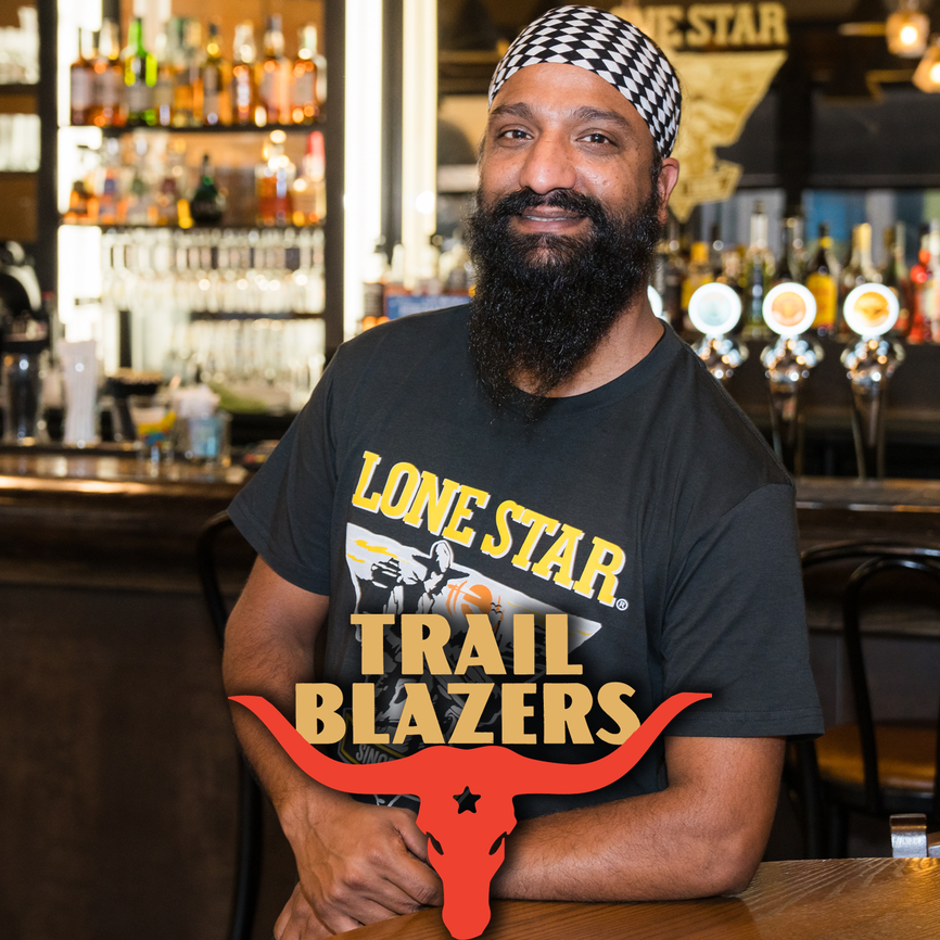 Smiling man with a beard and checkered headband stands at a bar, wearing a "Lone Star Trail Blazers" shirt.