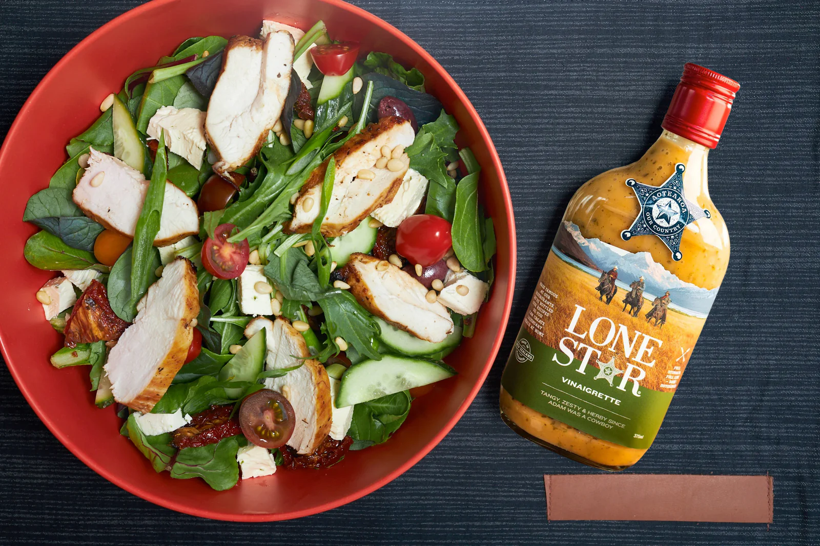 A vibrant salad with grilled chicken, spinach, tomatoes, cucumbers, and feta in a red bowl, next to a bottle of Lone Star vinaigrette.