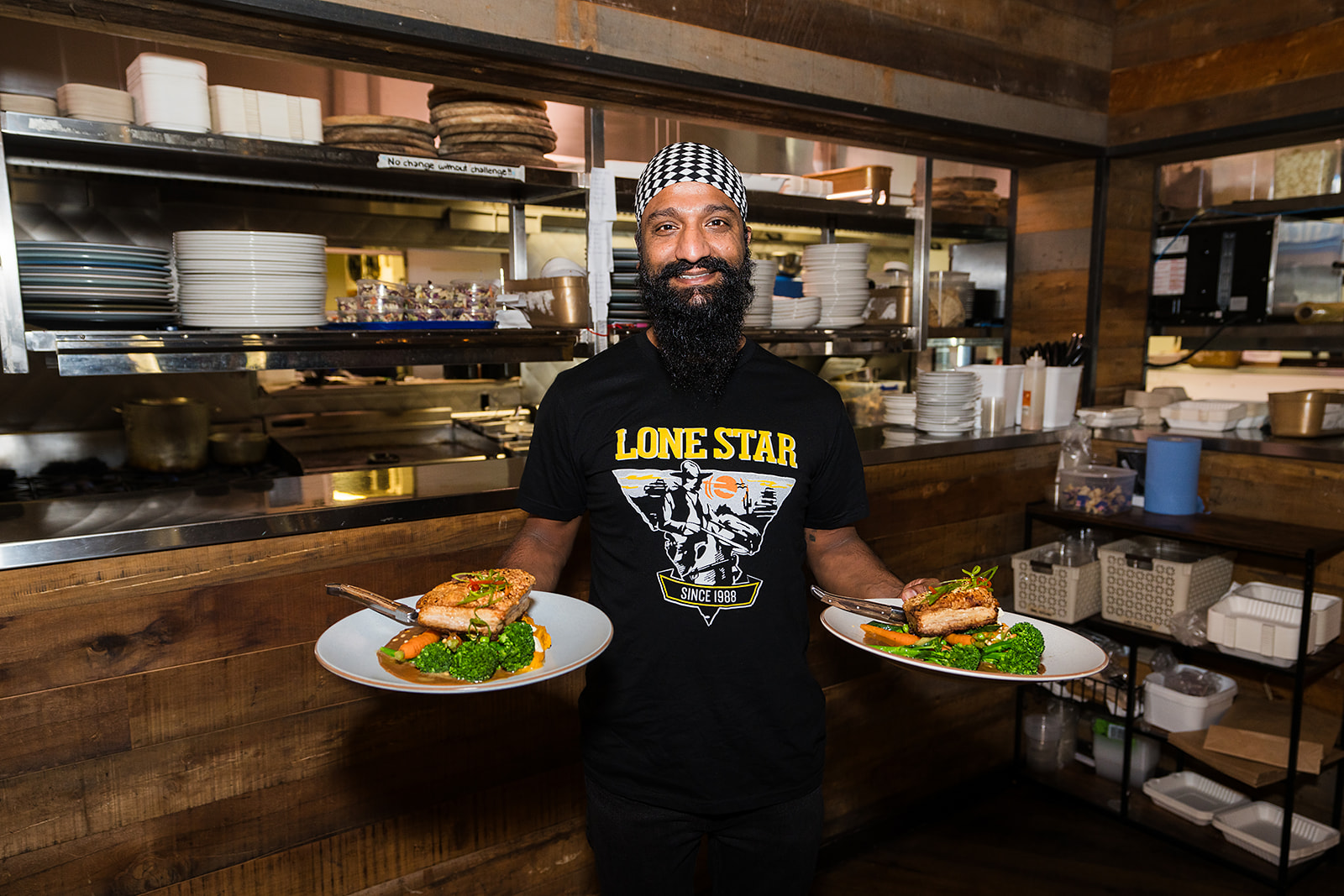 Man in a Lone Star shirt holding two plates of grilled chicken and vegetables in a rustic restaurant kitchen.