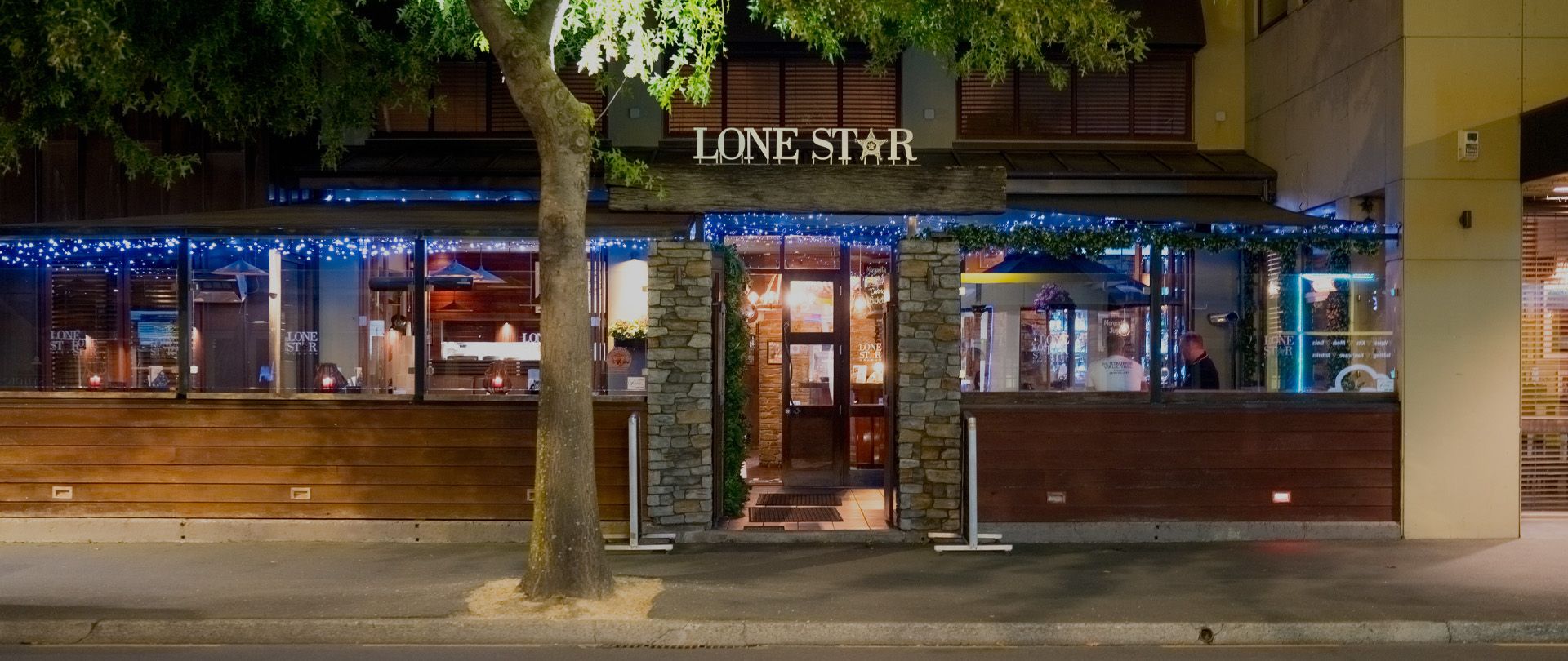 Exterior of a Lone Star restaurant at night, warmly lit with string lights, wooden and stone facade, and a tree in front.