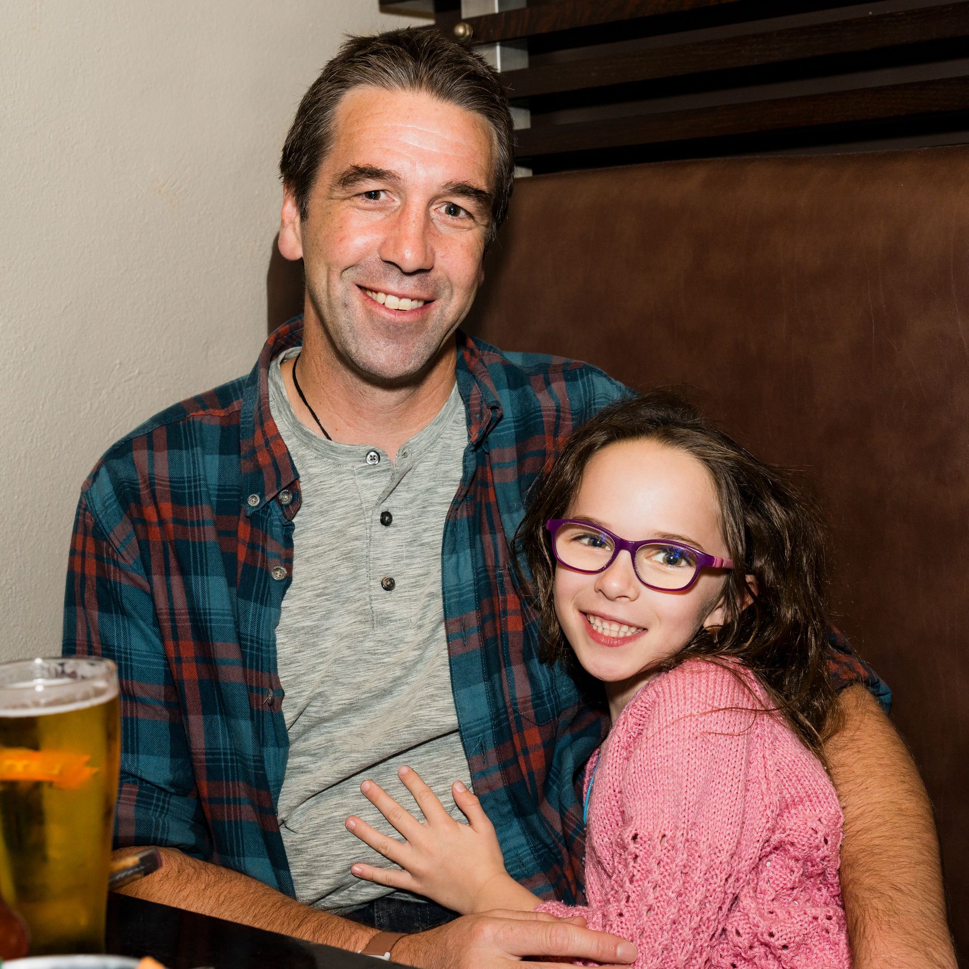A man and a young girl with glasses smile while sitting together in a booth at a restaurant. A glass of beer is on the table.