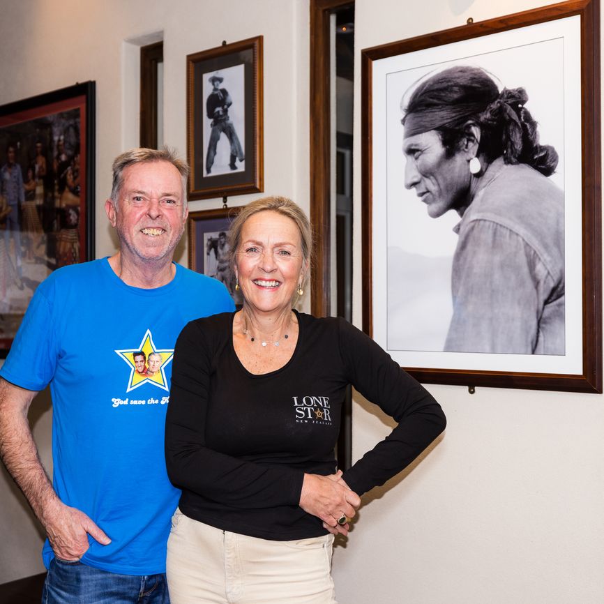 A smiling man and woman stand in front of framed photos on a wall, including a large black-and-white portrait.