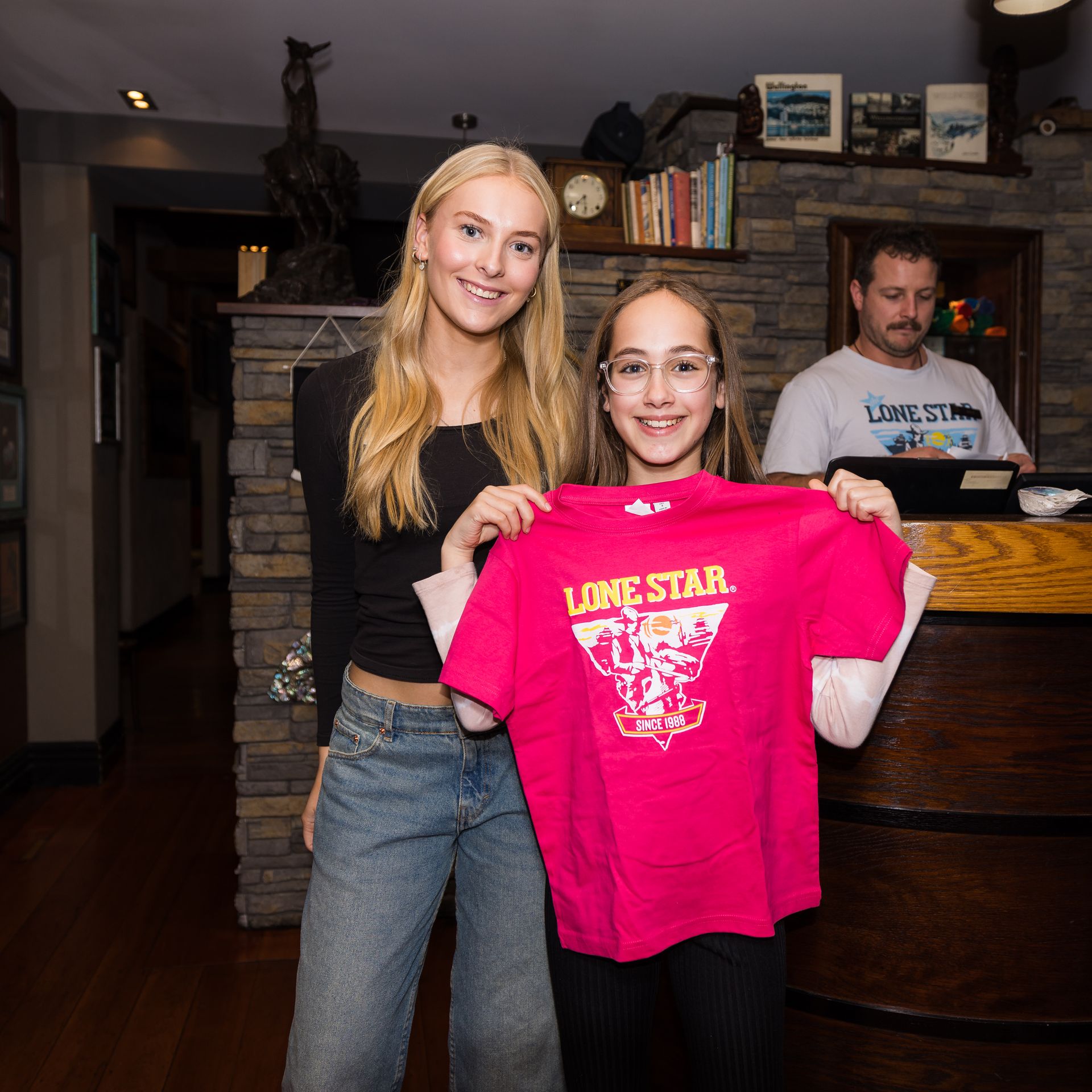 Two people smiling, one holding a pink "Lone Star" shirt in a cozy room with wooden floors and stone walls. A man stands behind a counter.