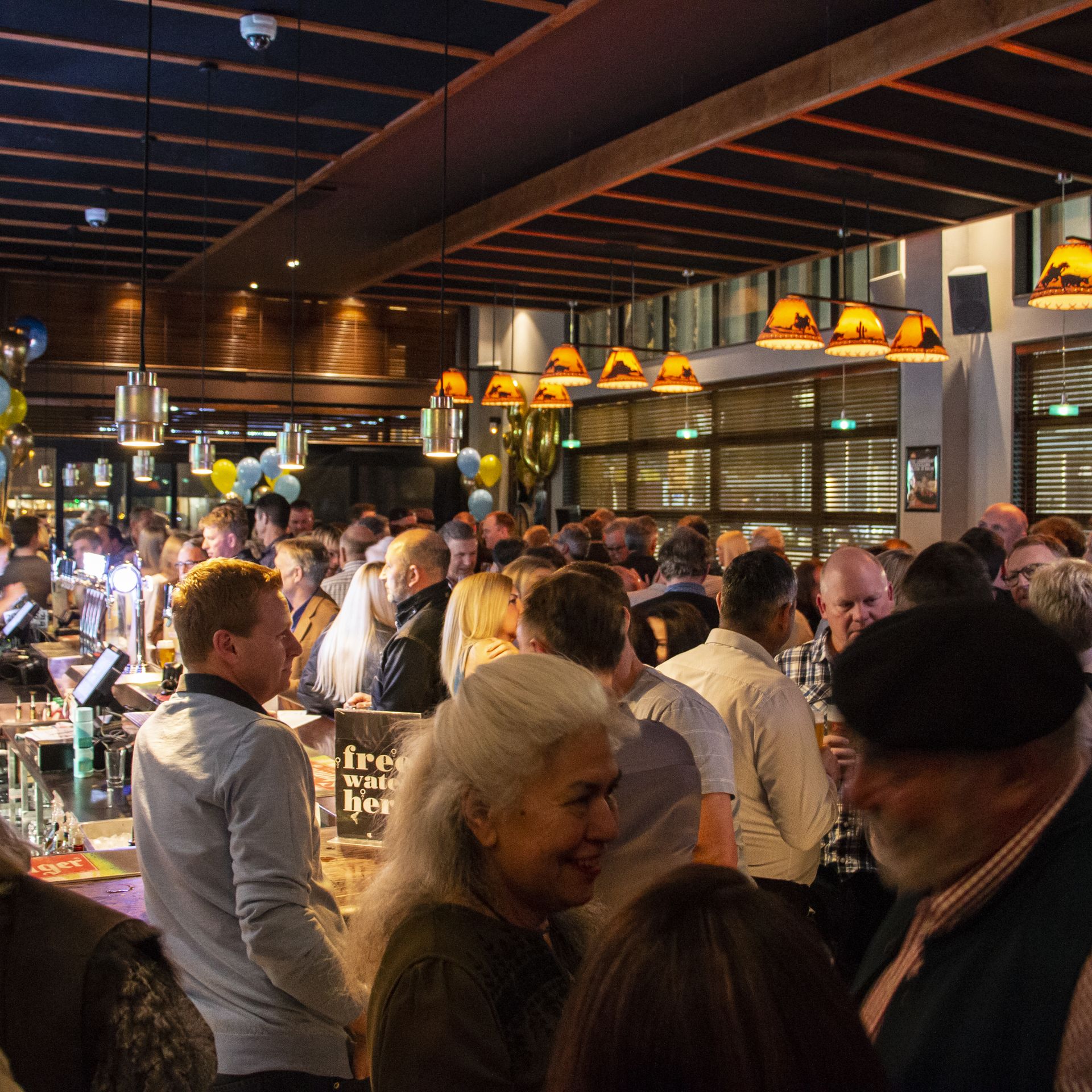 A crowded bar scene with people socialising, balloons, and warm lighting creating a lively atmosphere.