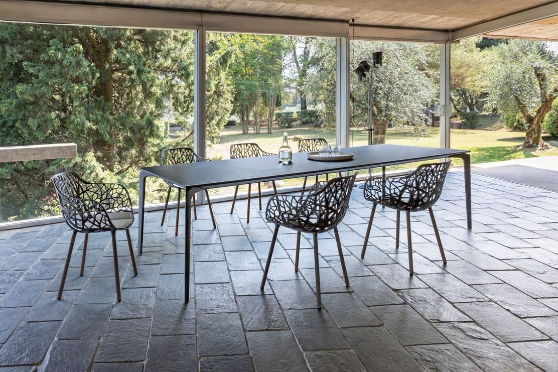 Glass-walled patio with a black rectangular dining table, six lattice-pattern chairs on stone tile floor overlooking a leafy garden.