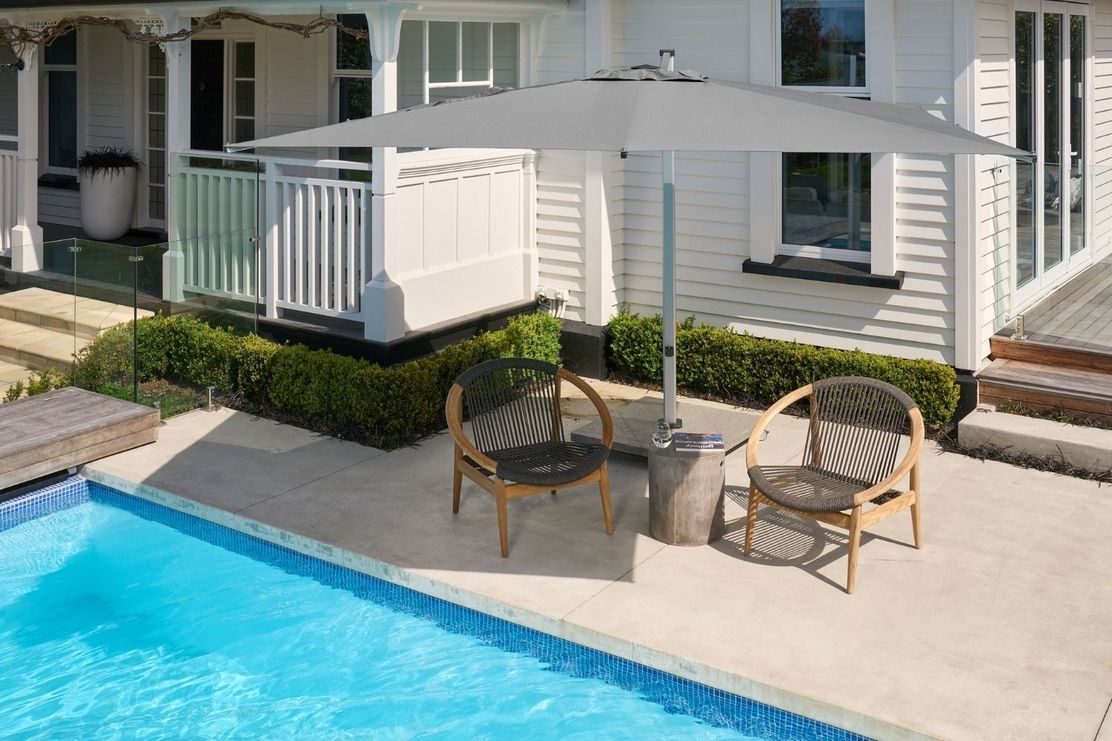 Poolside area with two Vincent Sheppard Frida Outdoor Lounge Chairs with Portico Dakota Outdoor Stool between, under the Jardinico Caractere 301 Outdoor Umbrella in Charcoal, next to a clear blue swimming pool against a white house exterior.