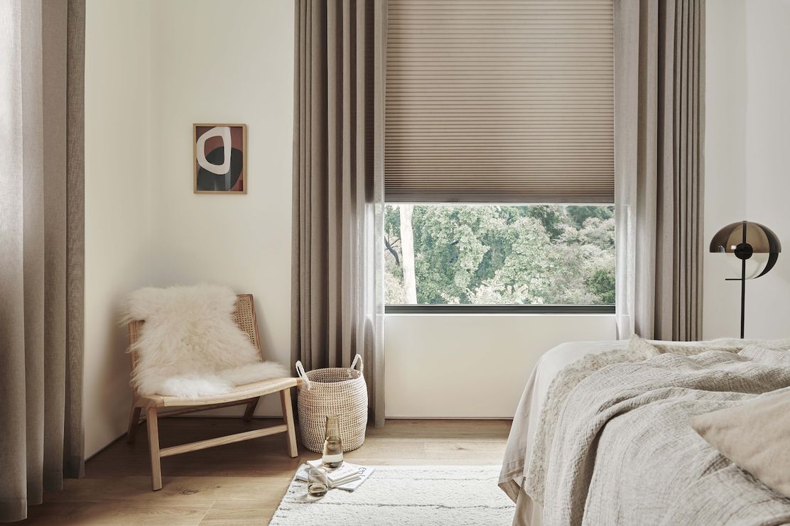 Cozy bedroom featuring a large window dressed in beige blinds and light, airy curtains, softening the view of greenery beyond.